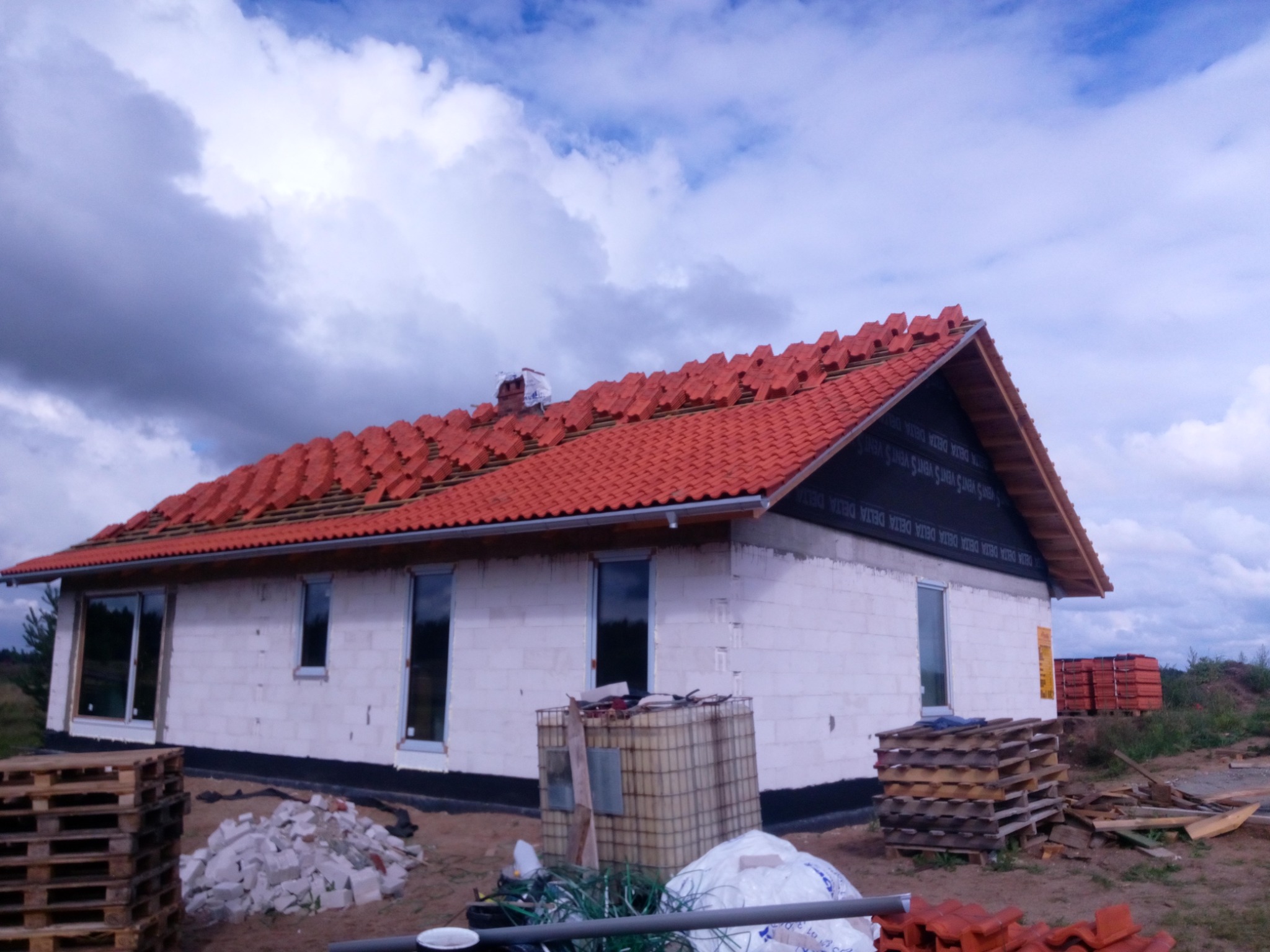 Dom w trakcie układania dachówki ceramicznej koloru ceglastego, widoczne palety z dachówką, niezabezpieczona ściana szczytowa, białe ściany z bloczków, pochmurne niebo.