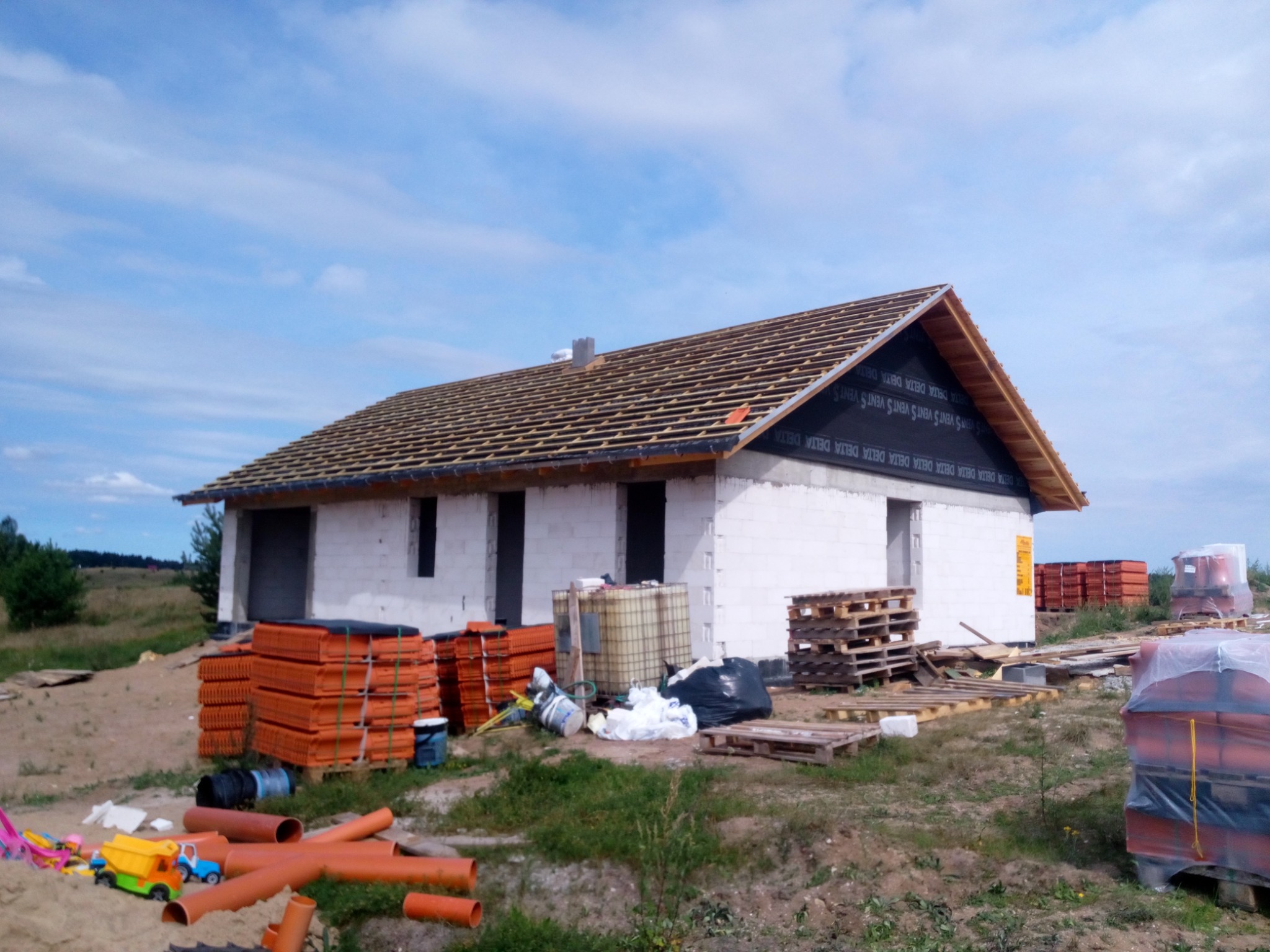 Dom w trakcie budowy z widocznym szkieletem dachu pokrytym membraną, przygotowane dachówki ułożone na paletach obok, porozrzucane rury kanalizacyjne i zabawki dziecięce na pierwszym planie.