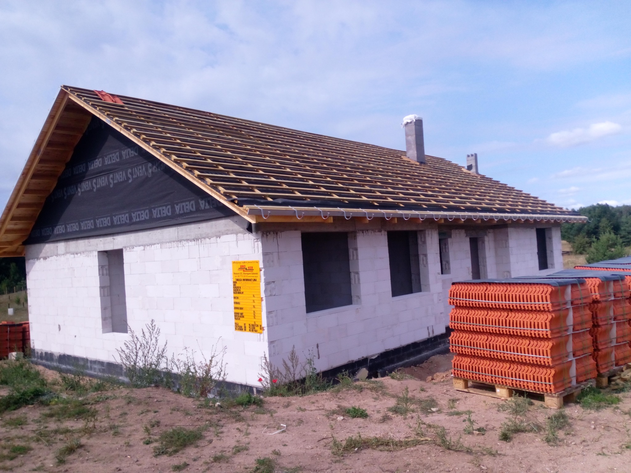 Budowa domu jednorodzinnego: widok na szkielet dachu z membraną, przygotowane łaty, palety z dachówką ceramiczną obok budynku z bloczków.