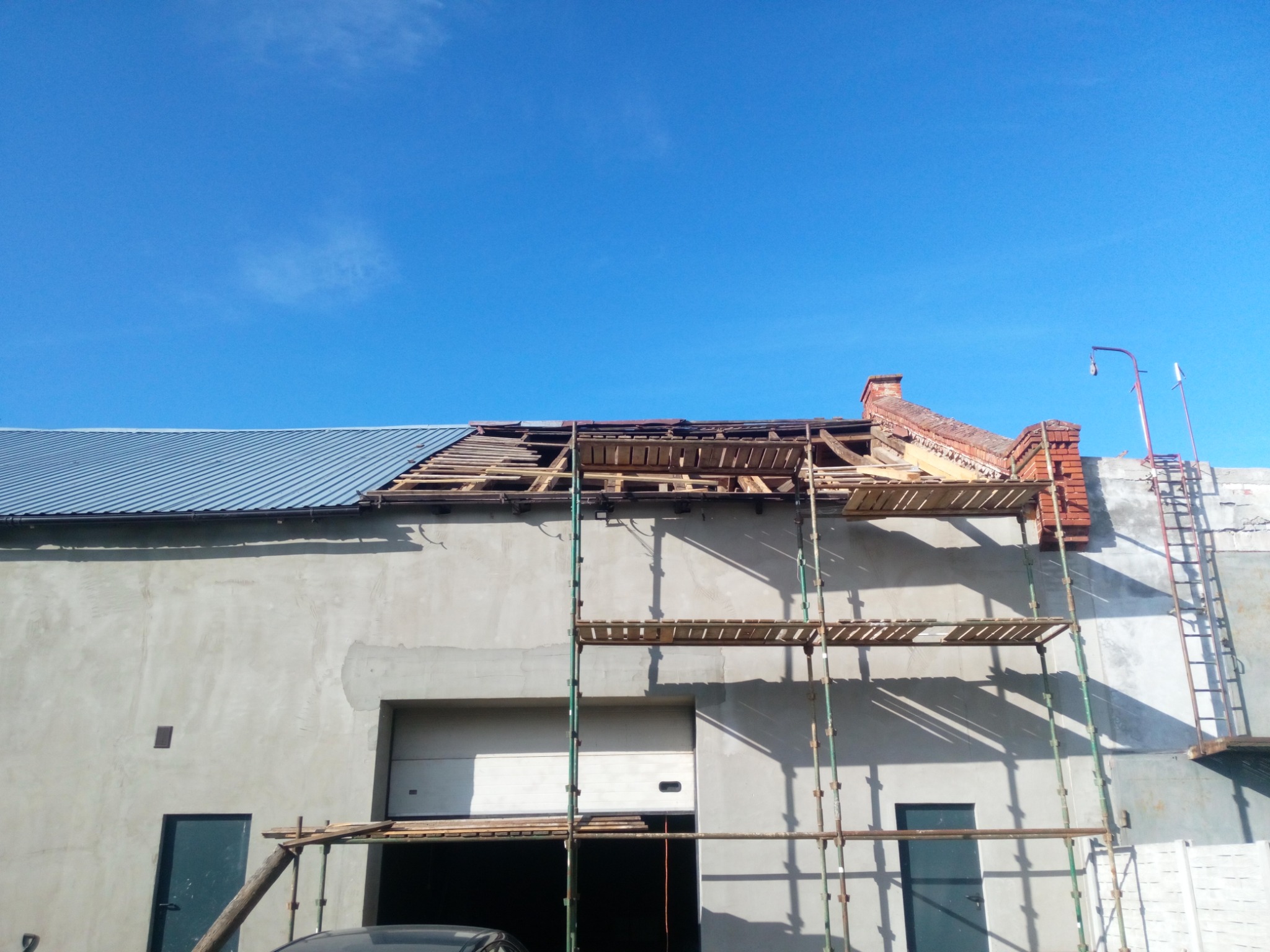 Budynek z częściowo zdjętym poszyciem dachowym, widoczne elementy konstrukcji dachu, rusztowanie i komin z czerwonej cegły na tle błękitnego nieba.