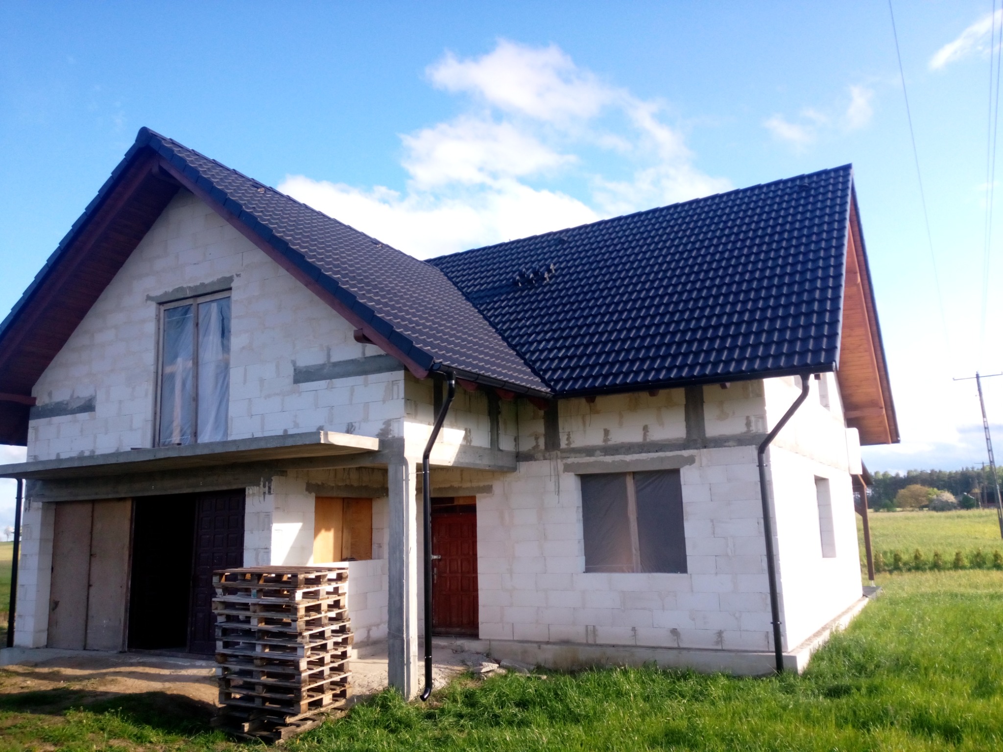 Niedokończony dom z czarną dachówką, widoczne rynny, okna zabezpieczone folią, palety przed garażem, trawa i błękitne niebo w tle.