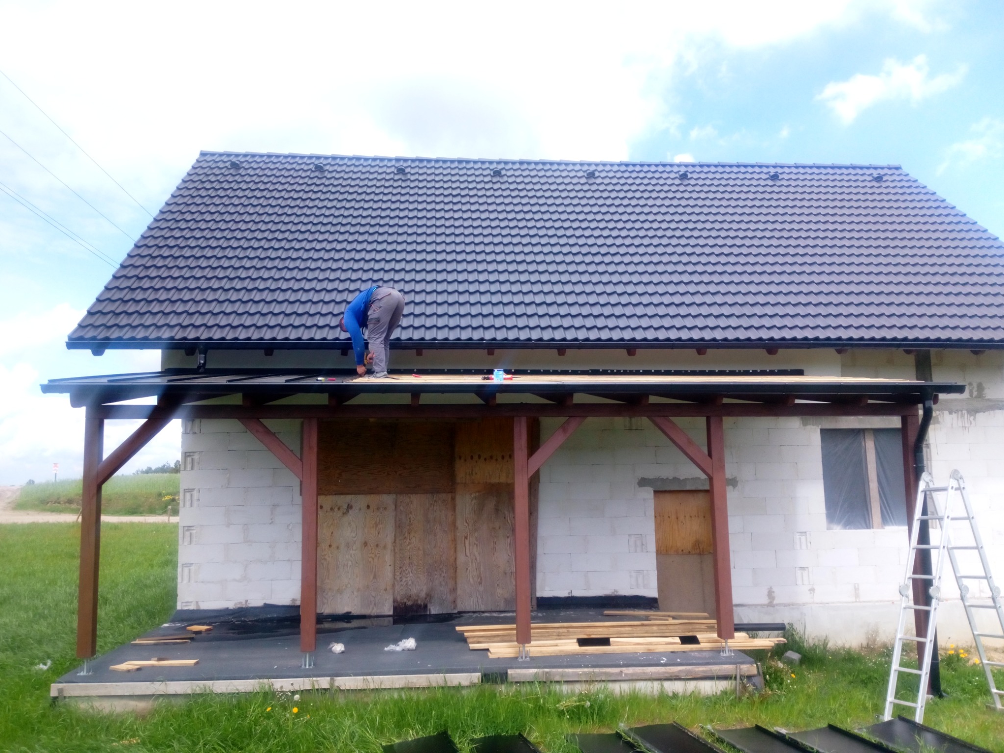 Dekarz montuje czarne dachówki na nowym dachu domu z białego bloczka, widoczne narzędzia i fragment konstrukcji werandy.