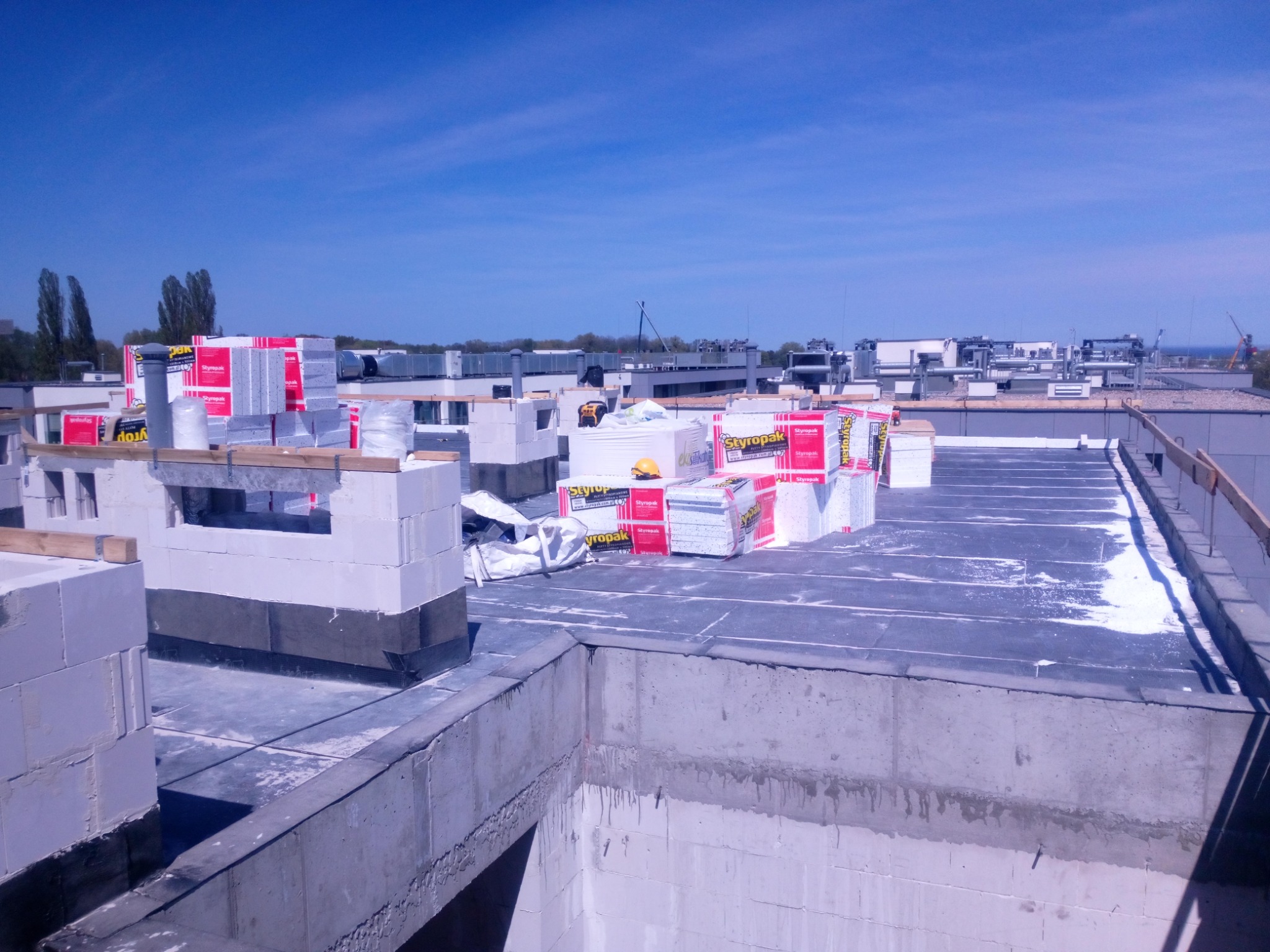 Płaski dach budynku w trakcie ocieplania, widoczne bloczki betonowe, izolacja termiczna w postaci styropianu w paczkach oraz pokrycie papą.