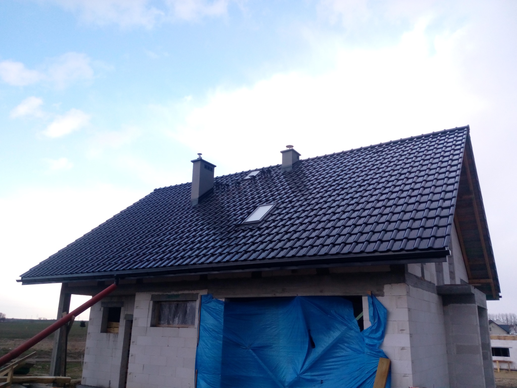 Nowy, czarny dach z dachówki ceramicznej na budynku w trakcie budowy, widoczne kominy i okna dachowe, część elewacji zakryta niebieską plandeką.