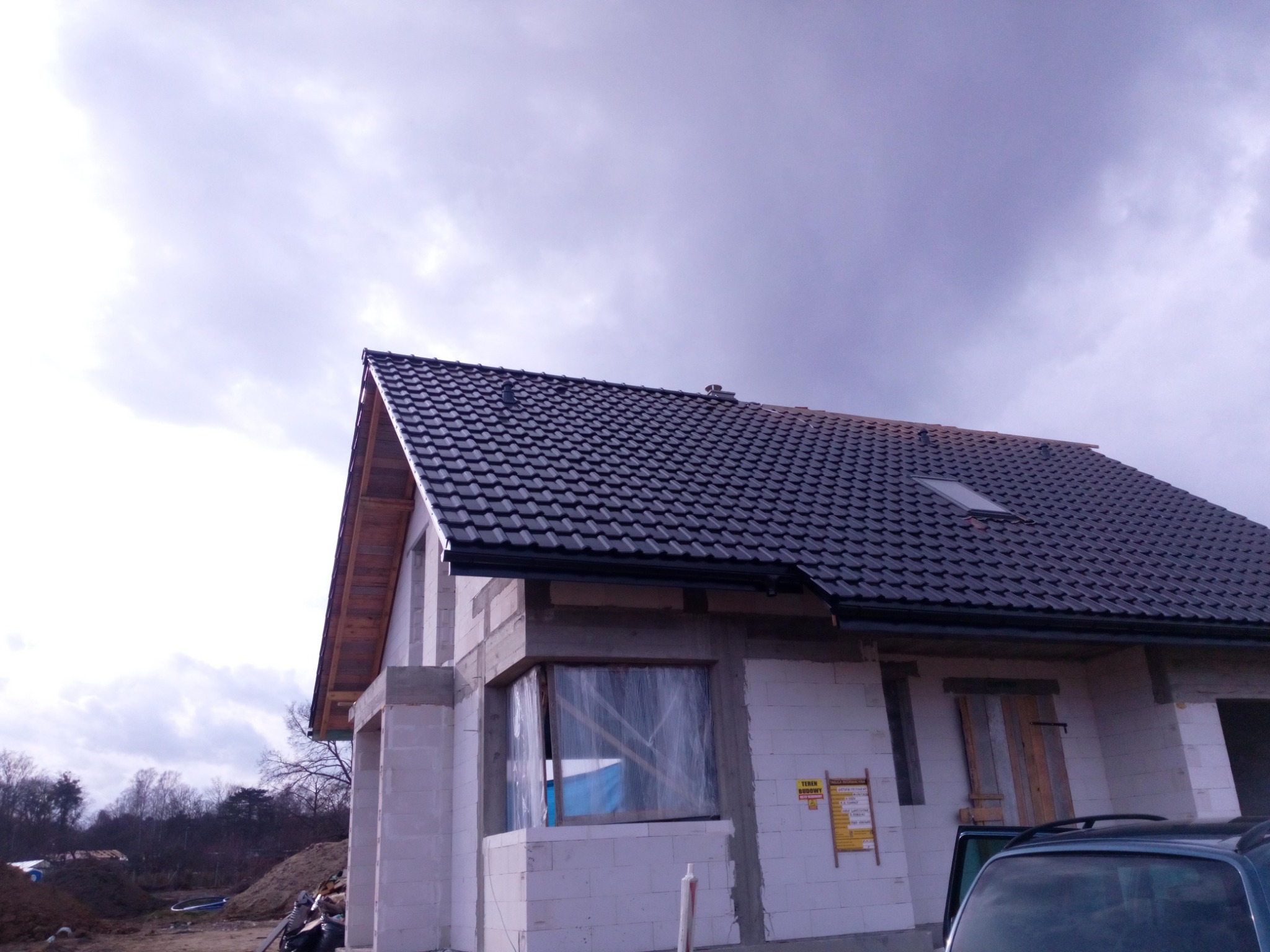 Dom w trakcie budowy z nowym, ciemnym dachem z dachówki ceramicznej i zamontowanymi rynnami, widoczne okno zabezpieczone folią i teren budowy.