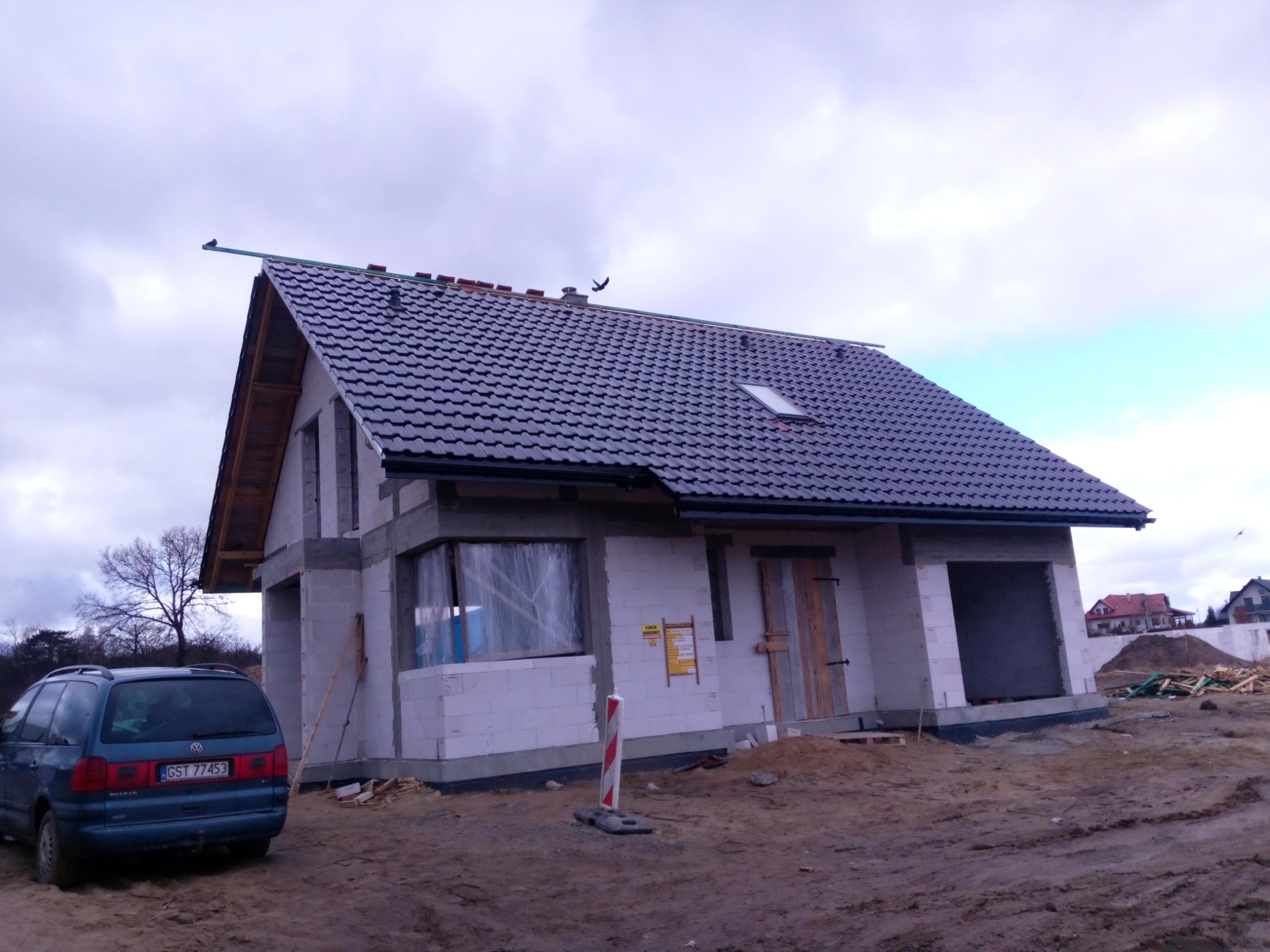 Dom w trakcie budowy z szarą dachówką, widoczne okno dachowe, elementy drewniane konstrukcji dachu, auto zaparkowane obok.
