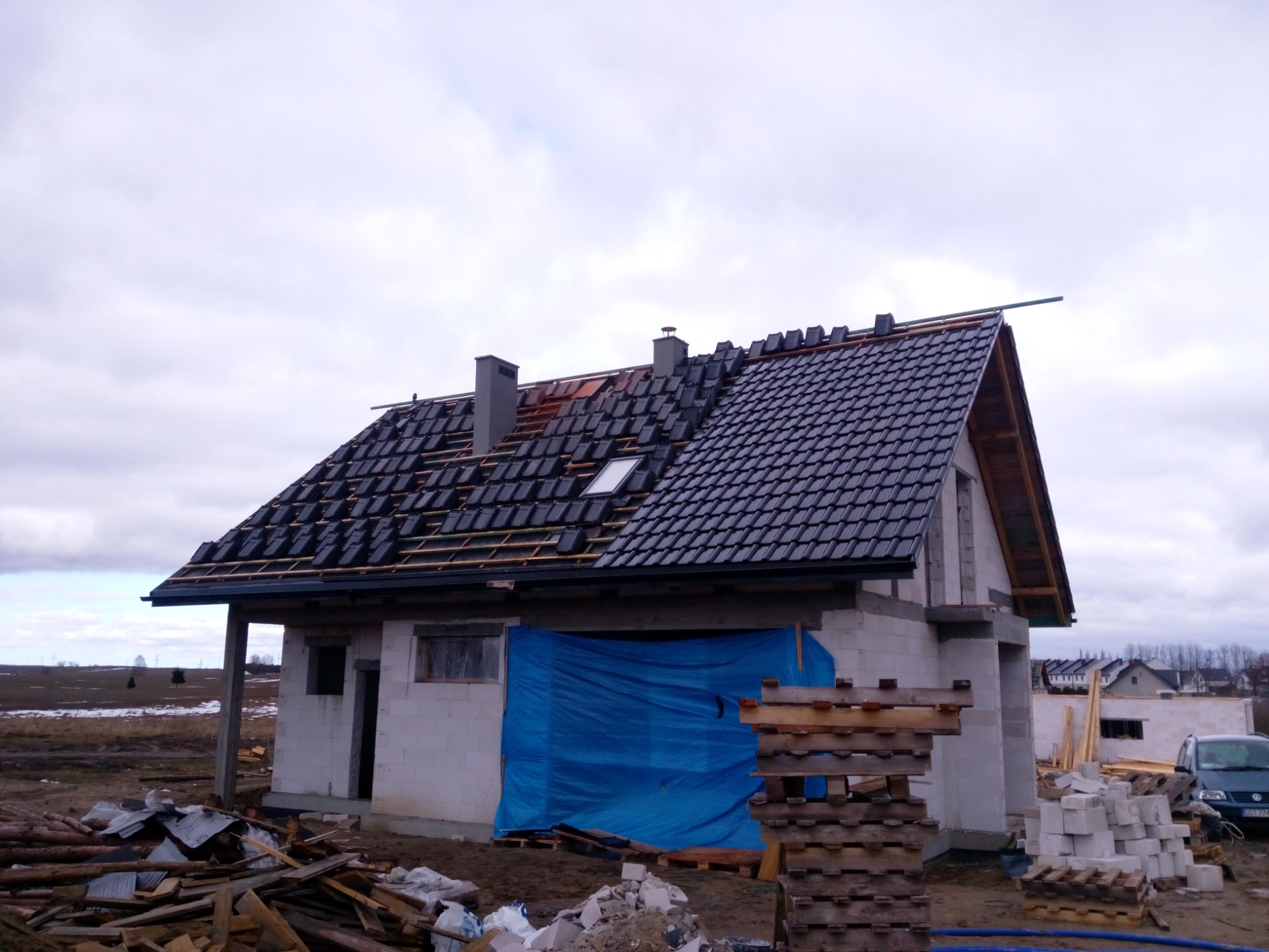 Dom w trakcie budowy z częściowo położonym, czarnym dachem z dachówki ceramicznej, zabezpieczony niebieską plandeką, z widocznymi elementami drewnianej konstrukcji i stosem materiałów budowlanych.