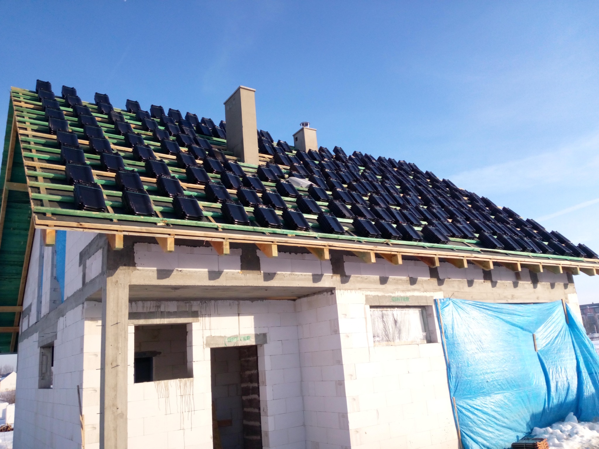 Budowa domu jednorodzinnego w trakcie. Widoczny szkielet dachu z częściowo ułożoną, czarną dachówką ceramiczną, kominy oraz ściany z białego bloczka. Część okien zabezpieczona folią.