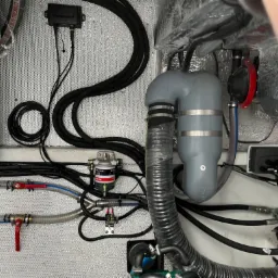 Kompleksowy system rur i przewodów wewnątrz łodzi, z widocznym filtrem paliwa Delphi, szarym separatorem wody, zaworami z czerwonymi uchwytami i czarną skrzynką Garmin na perforowanej ścianie.