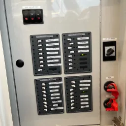 Szary panel elektryczny z bezpiecznikami i przełącznikami na jachcie, widoczne wskaźniki napięcia LED, opisane obwody: gniazda, klimatyzacja, oświetlenie, pompy i urządzenia sanitarne.