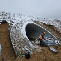 Budowa przepustu drogowego z kamiennym obłożeniem i betonowymi schodami w zimowej scenerii; pracownik w odblaskowej kamizelce przygotowuje kostkę brukową.