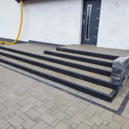 Wyłożony kostką brukową podjazd z czterostopniowymi schodami prowadzącymi do wejścia domu, wykończony szarą kostką granitową, widoczna żółta rura odprowadzająca wodę z dachu i pracownik w tle.