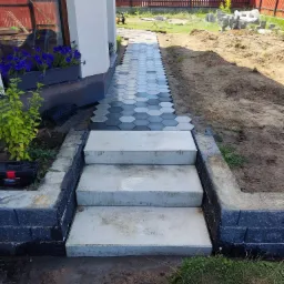 Trzy betonowe stopnie prowadzące do ścieżki z kostki w kształcie szarego i białego sześciokąta, obrzeża z ciemnych bloczków, w tle czerwone ogrodzenie i domy.