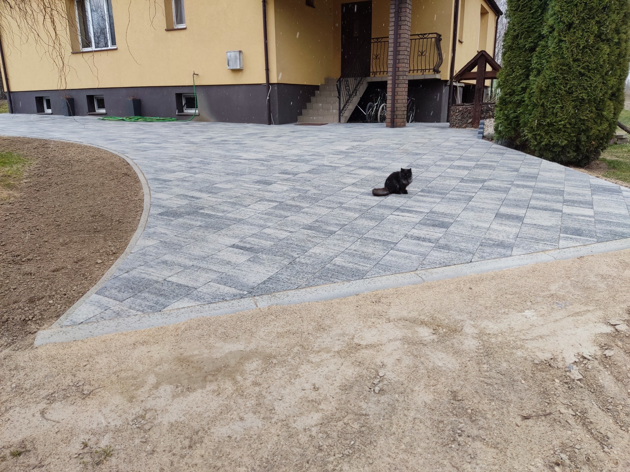 Nowo ułożony, szary chodnik z kostki brukowej prowadzący do domu, na którym siedzi czarny kot. W tle widoczne schody wejściowe i elementy architektury ogrodowej.