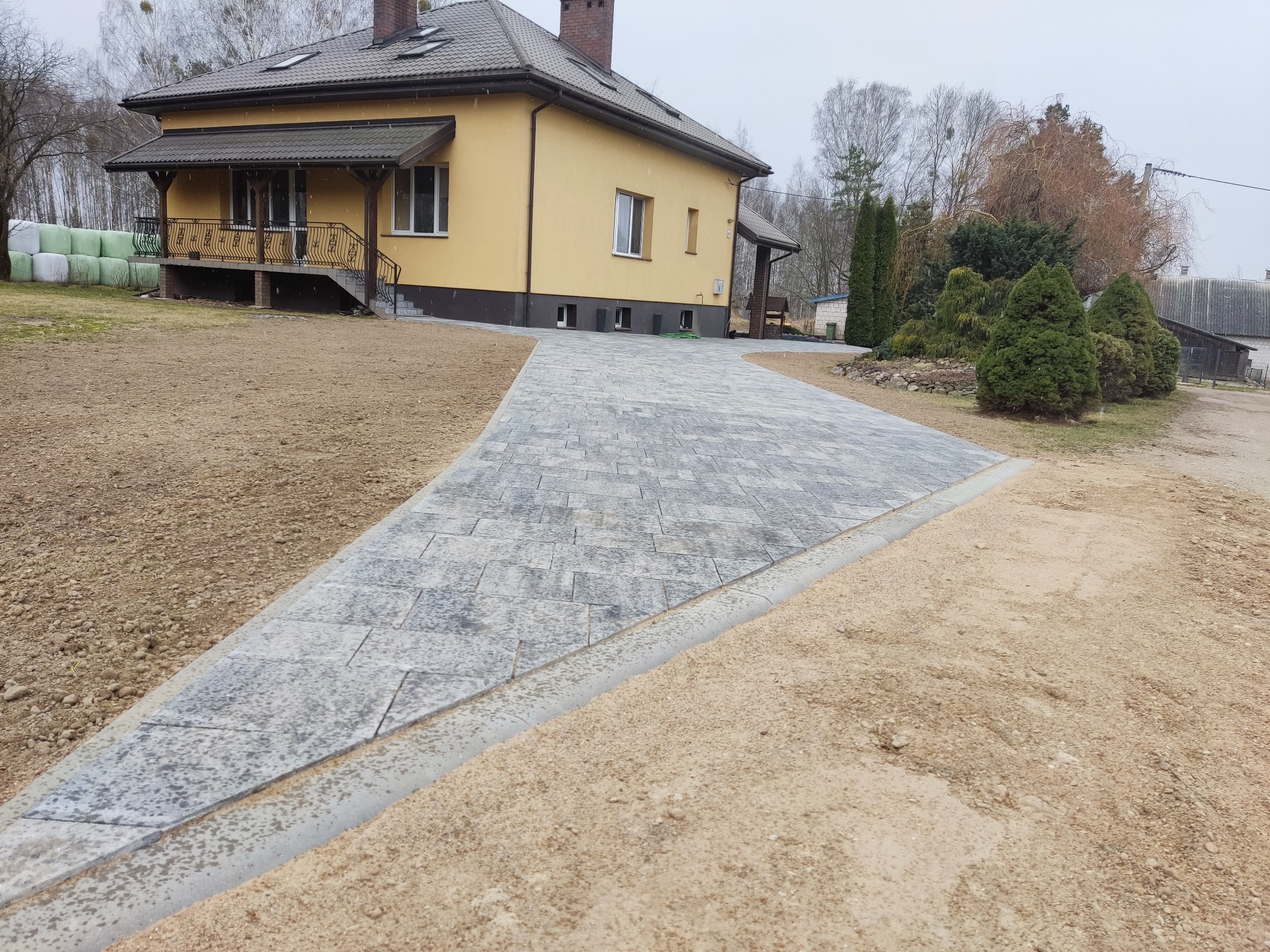 Szeroki podjazd z szarej kostki brukowej prowadzący do żółtego domu z ciemnobrązowym dachem, widoczne obrzeża podjazdu i przygotowane podłoże z piasku.