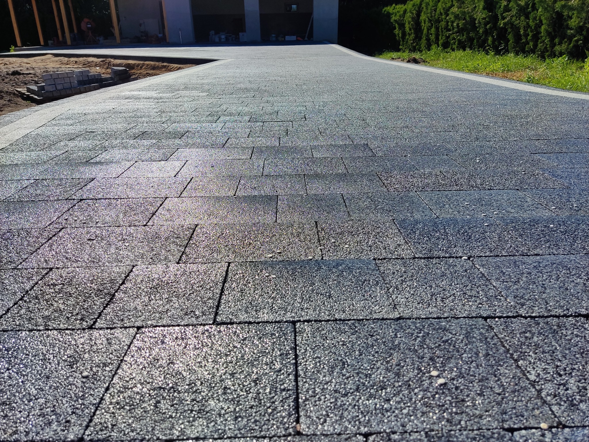 Nowo ułożona, ciemnoszara nawierzchnia z kostki brukowej, z widocznym fragmentem posesji w tle i stertą kostki przygotowanej do dalszych prac.