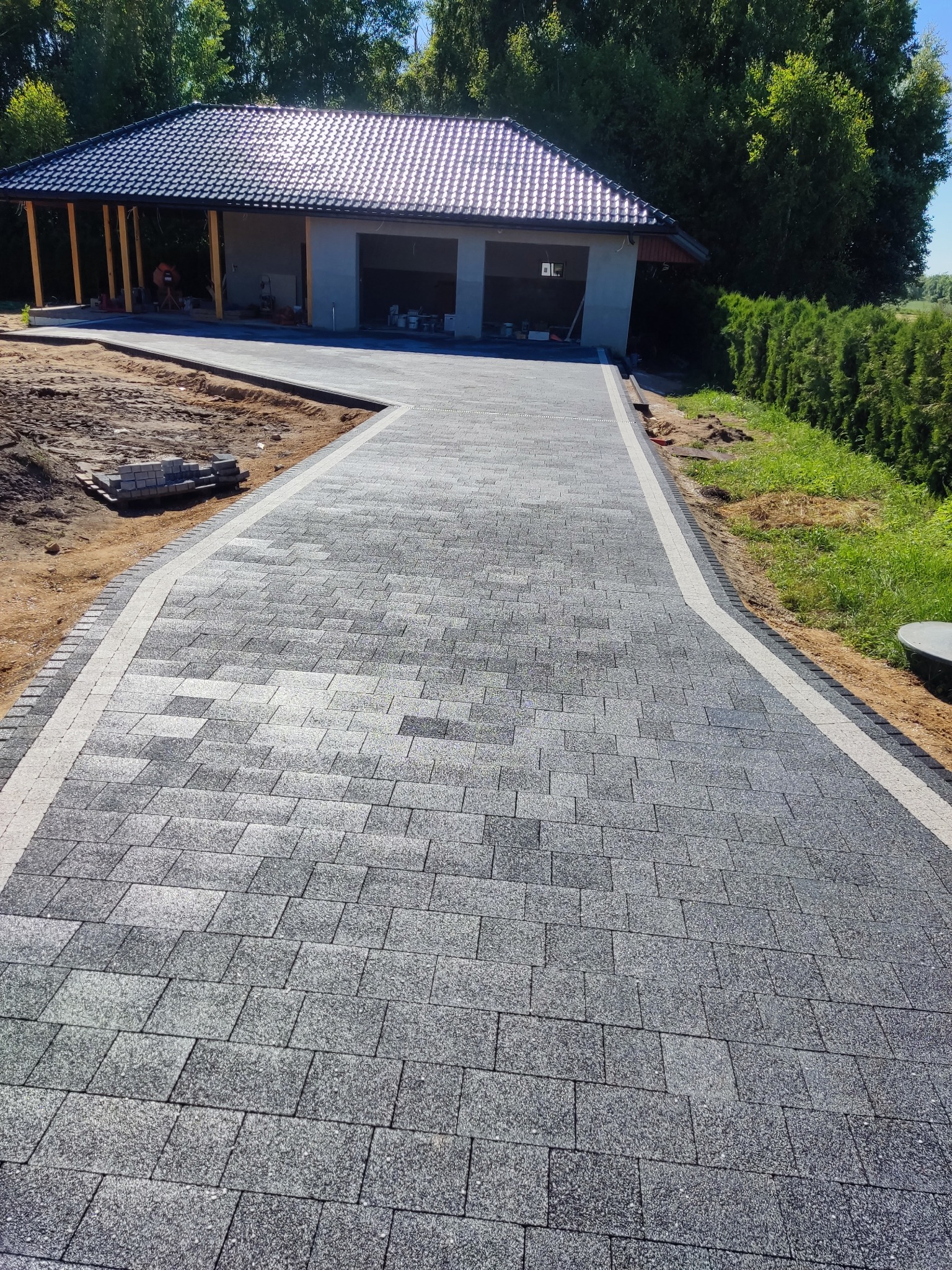 Długa, zakrzywiona ścieżka z szarej kostki brukowej prowadząca do garażu z zadaszeniem, obrzeża wykończone jaśniejszą kostką, widoczne zapasy kostki brukowej.