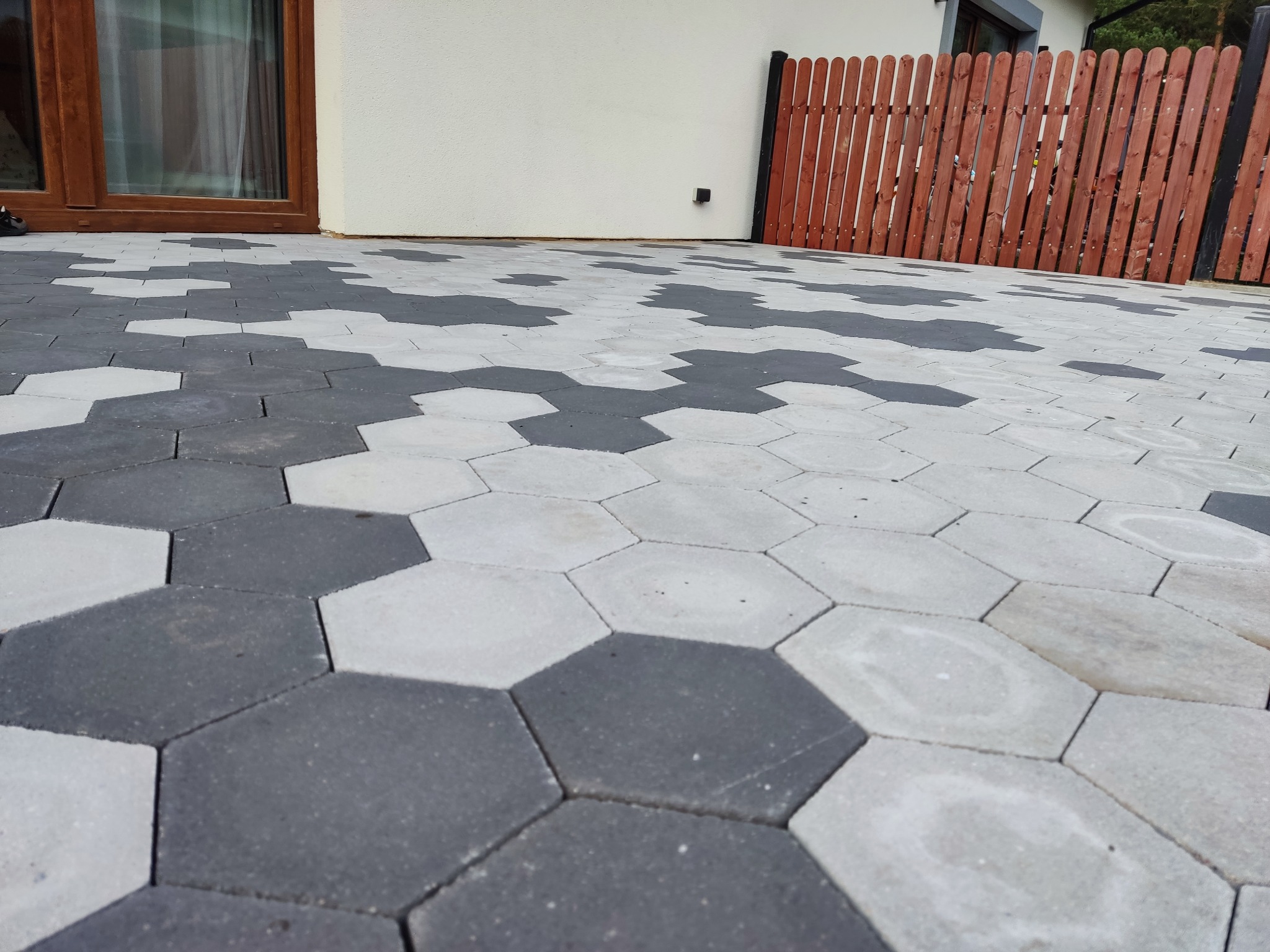 Ułożona nawierzchnia z sześciokątnej kostki w odcieniach szarości i bieli, tworząca wzór przed budynkiem z brązowymi ramami okiennymi i drewnianym płotem.