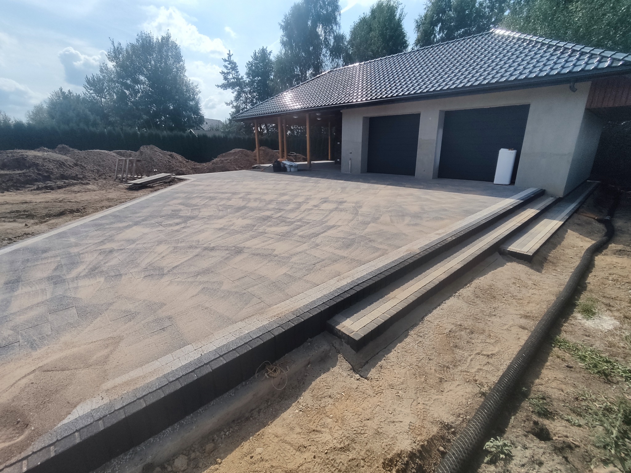 Nowo ułożona, szara kostka brukowa na podjeździe przed garażem z dwoma bramami. Widoczne obrzeża i schody z kostki, w tle stosy ziemi i drzewa.