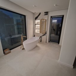 PPHU ADMAR MARTYNA KAŹMIERCZAK - Nowoczesna łazienka z wolnostojącą wanną, złotą armaturą i grzejnikiem drabinkowym na tle marmurowej ściany. Duże okno z widokiem na zewnątrz.