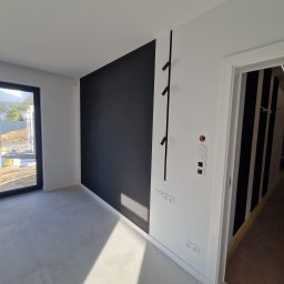 PPHU ADMAR MARTYNA KAŹMIERCZAK - Minimalistyczne wnętrze: czarno-biała ściana z wieszakiem, widoczne gniazdka i włączniki. W tle drzwi z pionowymi pasami i okno z widokiem na plac budowy.