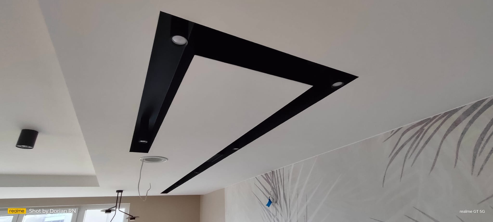 Nowoczesny sufit podwieszany z czarnymi elementami dekoracyjnymi i oświetleniem punktowym, widok z dołu, ściana z delikatnym wzorem roślinnym w tle.