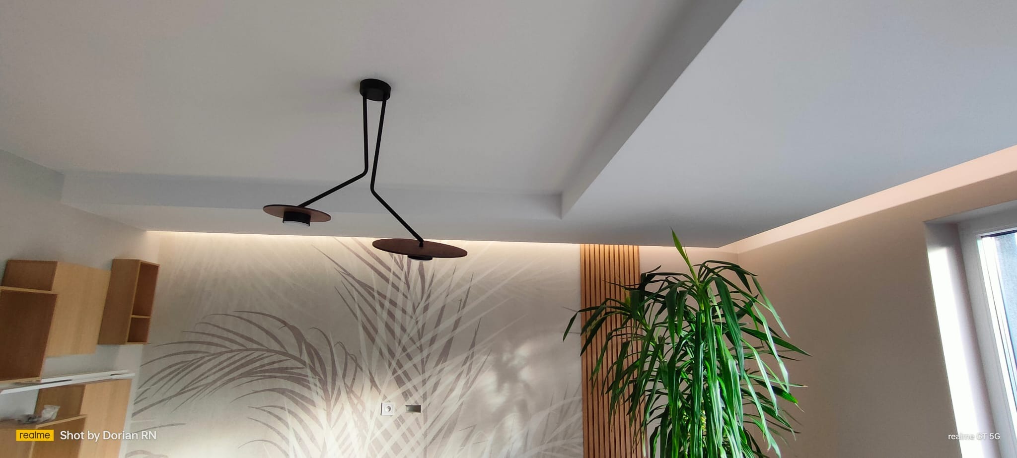 Nowoczesne wnętrze z podwieszanym sufitem, designerską lampą z dwoma okrągłymi kloszami, tapetą w palmy i wysoką rośliną doniczkową przy ścianie z drewnianymi panelami.