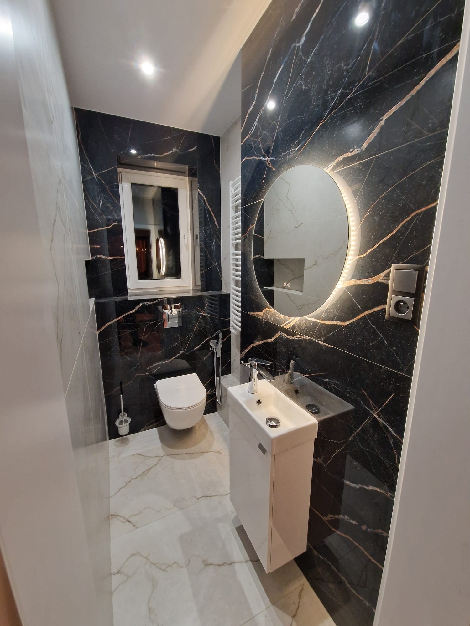 Elegancka łazienka z czarną marmurową ścianą akcentową, podświetlanym okrągłym lustrem, białą wiszącą toaletą i umywalką z szafką, wykończona płytkami podłogowymi imitującymi marmur.
