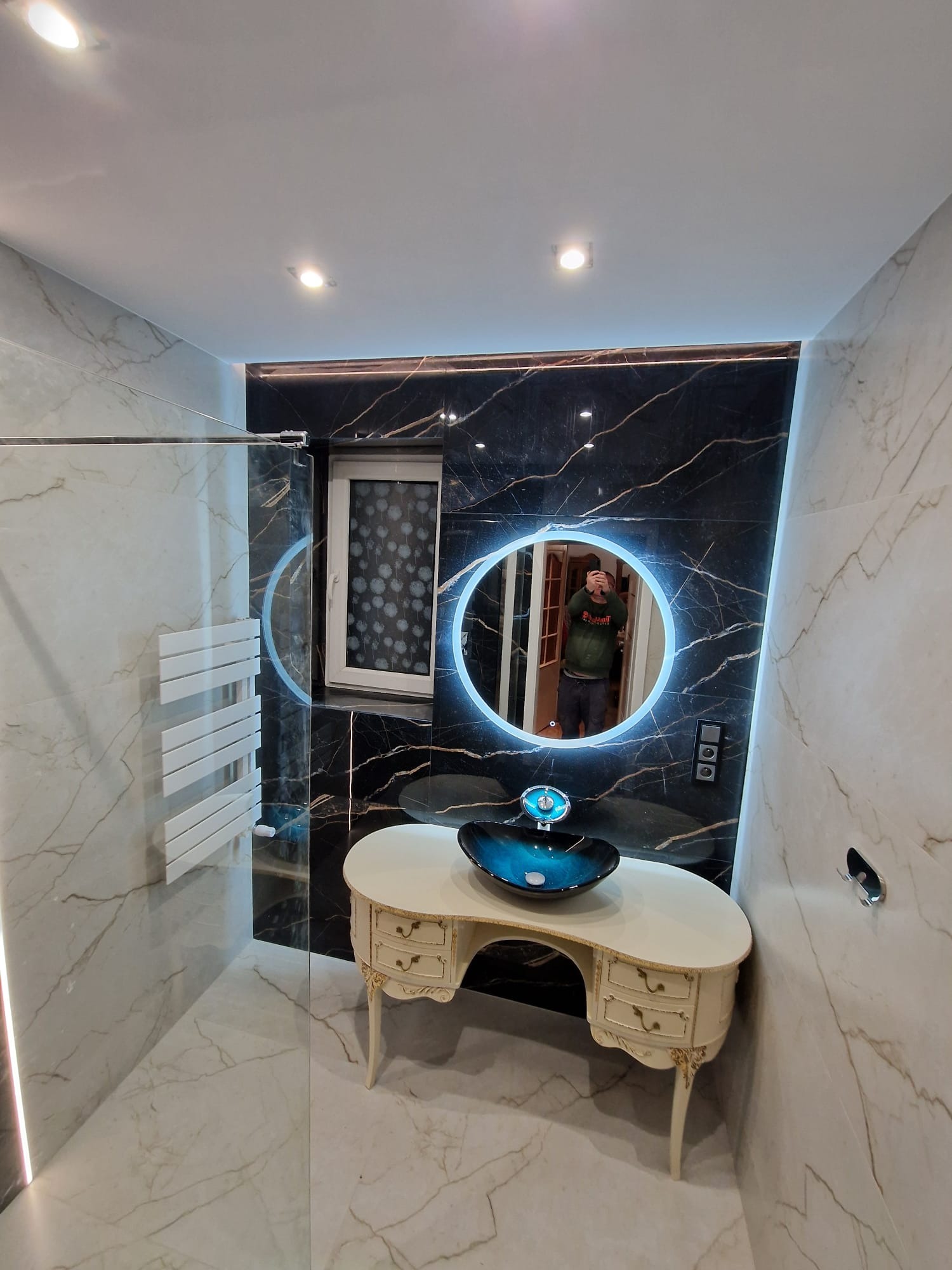 Elegancka łazienka z czarno-białymi marmurowymi płytkami, okrągłym lustrem z podświetleniem LED, szklaną kabiną prysznicową i toaletką w stylu retro z umywalką w odcieniach błękitu.