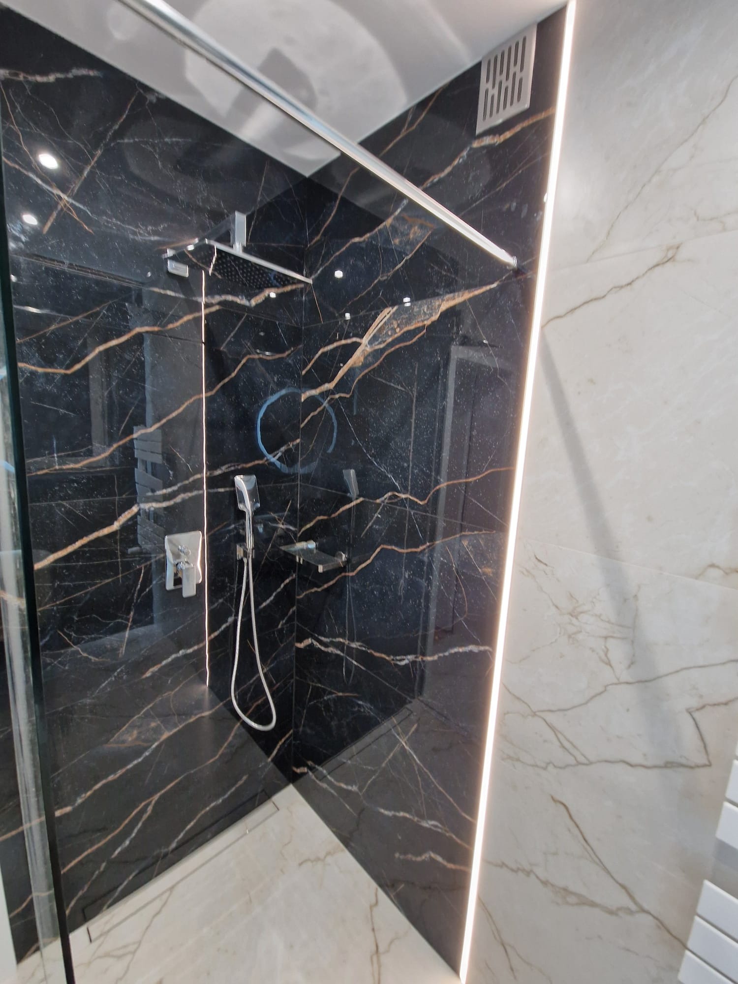 Nowoczesna kabina prysznicowa z czarnymi płytkami imitującymi marmur z złotymi żyłami, kontrastująca z jasnymi płytkami na sąsiedniej ścianie, oświetlona pionową listwą LED.