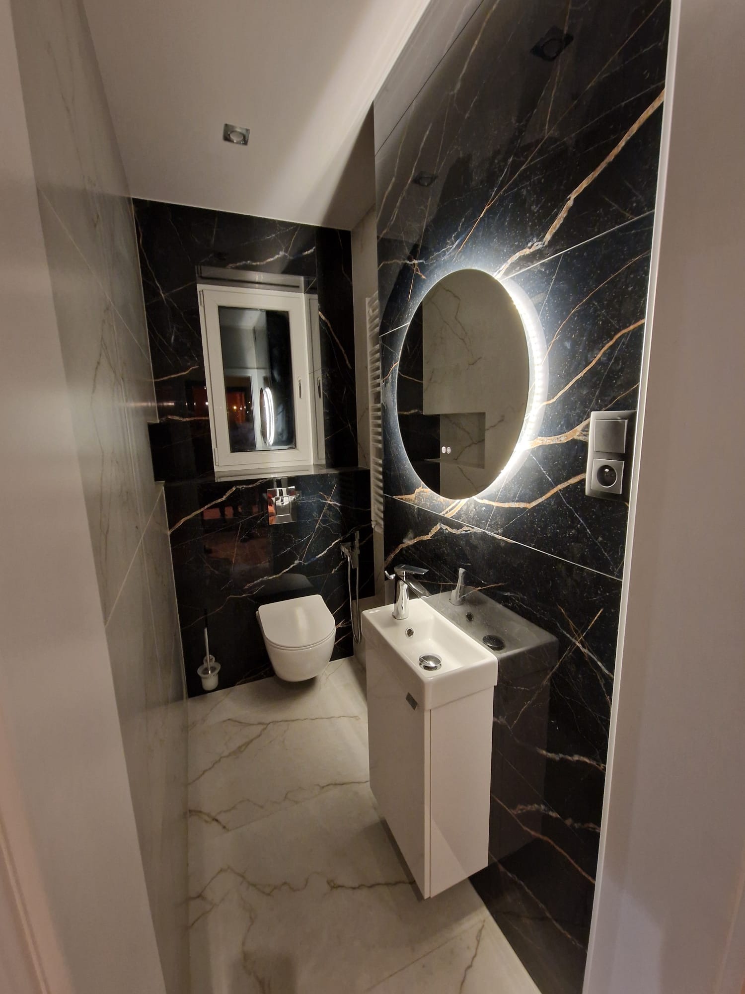 Nowoczesna łazienka z czarnymi marmurowymi płytkami, okrągłym lustrem z podświetleniem LED, białą umywalką na szafce i podwieszaną toaletą.