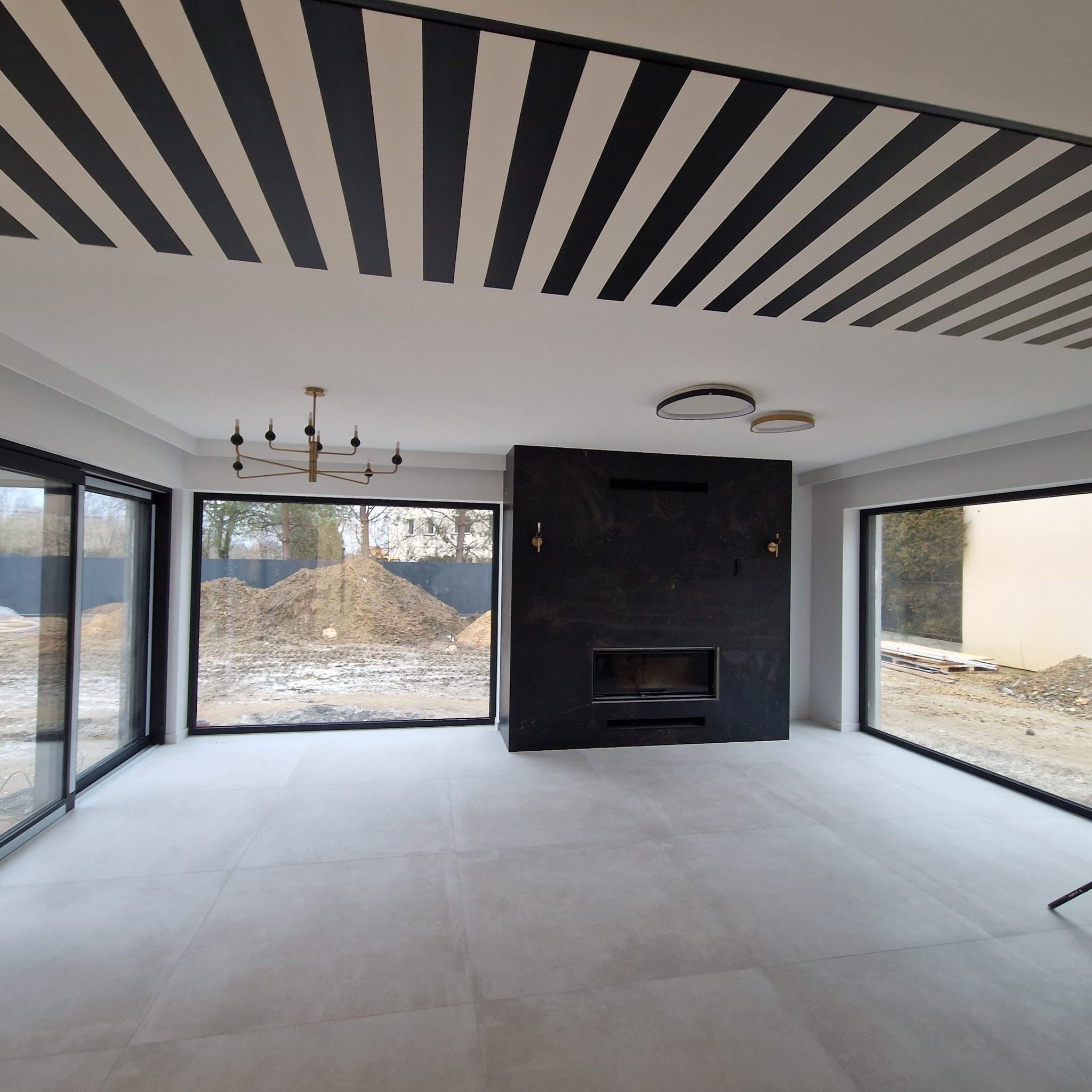 Nowoczesny salon z czarnym kominkiem i dużymi oknami. Sufit zdobią czarno-białe pasy. Widoczne minimalistyczne lampy i złoty żyrandol. Na zewnątrz widok na plac budowy.