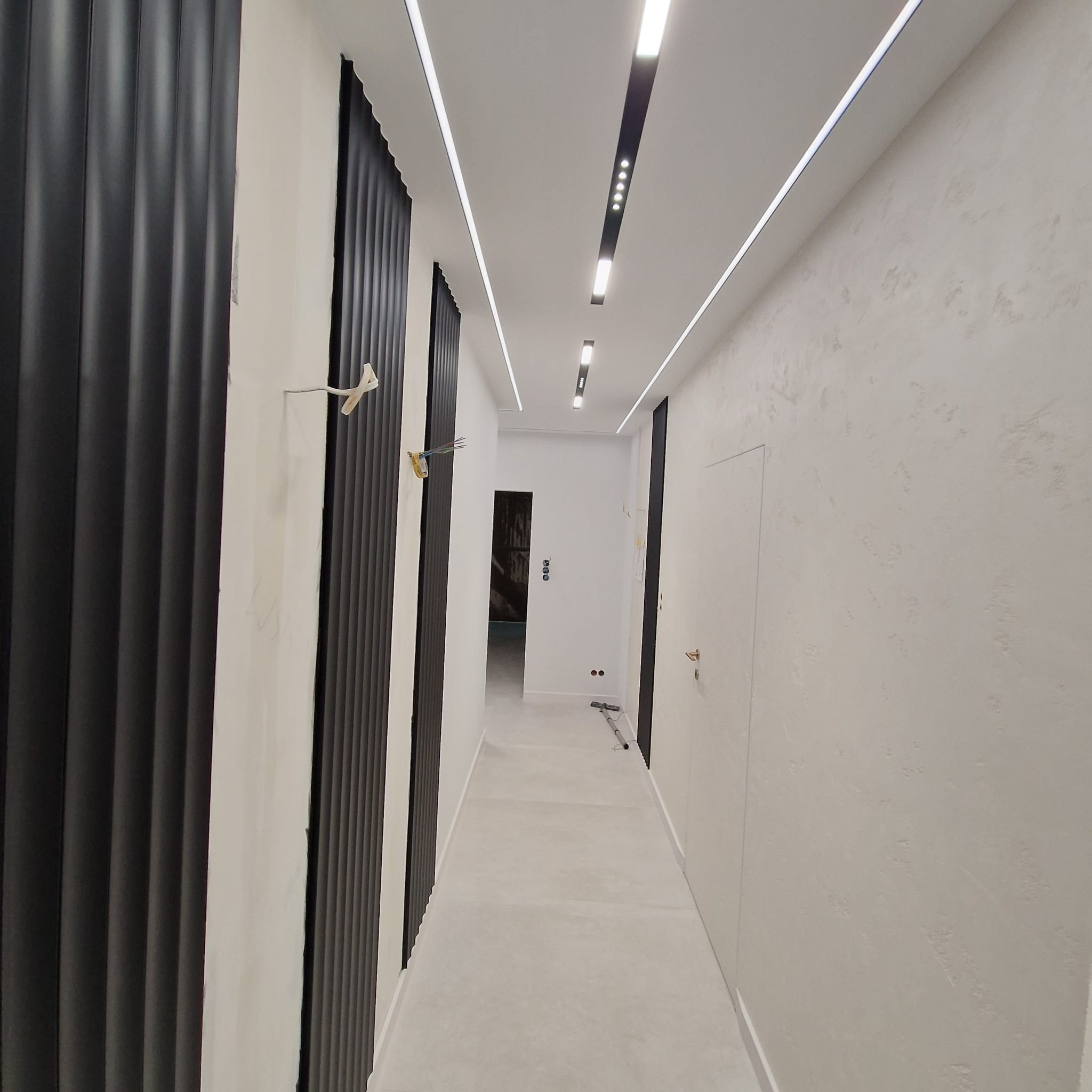 Nowoczesny korytarz po remoncie: białe ściany z fakturą, czarne panele dekoracyjne, oświetlenie liniowe LED. Minimalistyczny design, widok w głąb pomieszczenia.