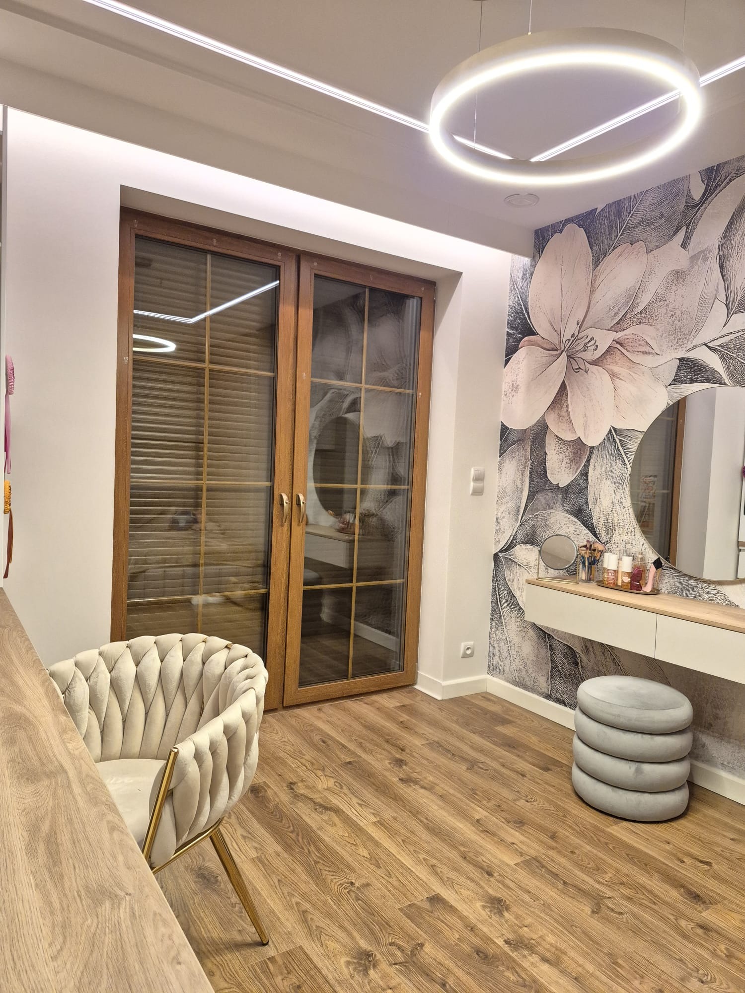 Elegancka toaletka z okrągłym lustrem i kwiatową tapetą, oświetlona nowoczesną lampą. Wnętrze dopełnia drewniana podłoga i stylowe krzesło.