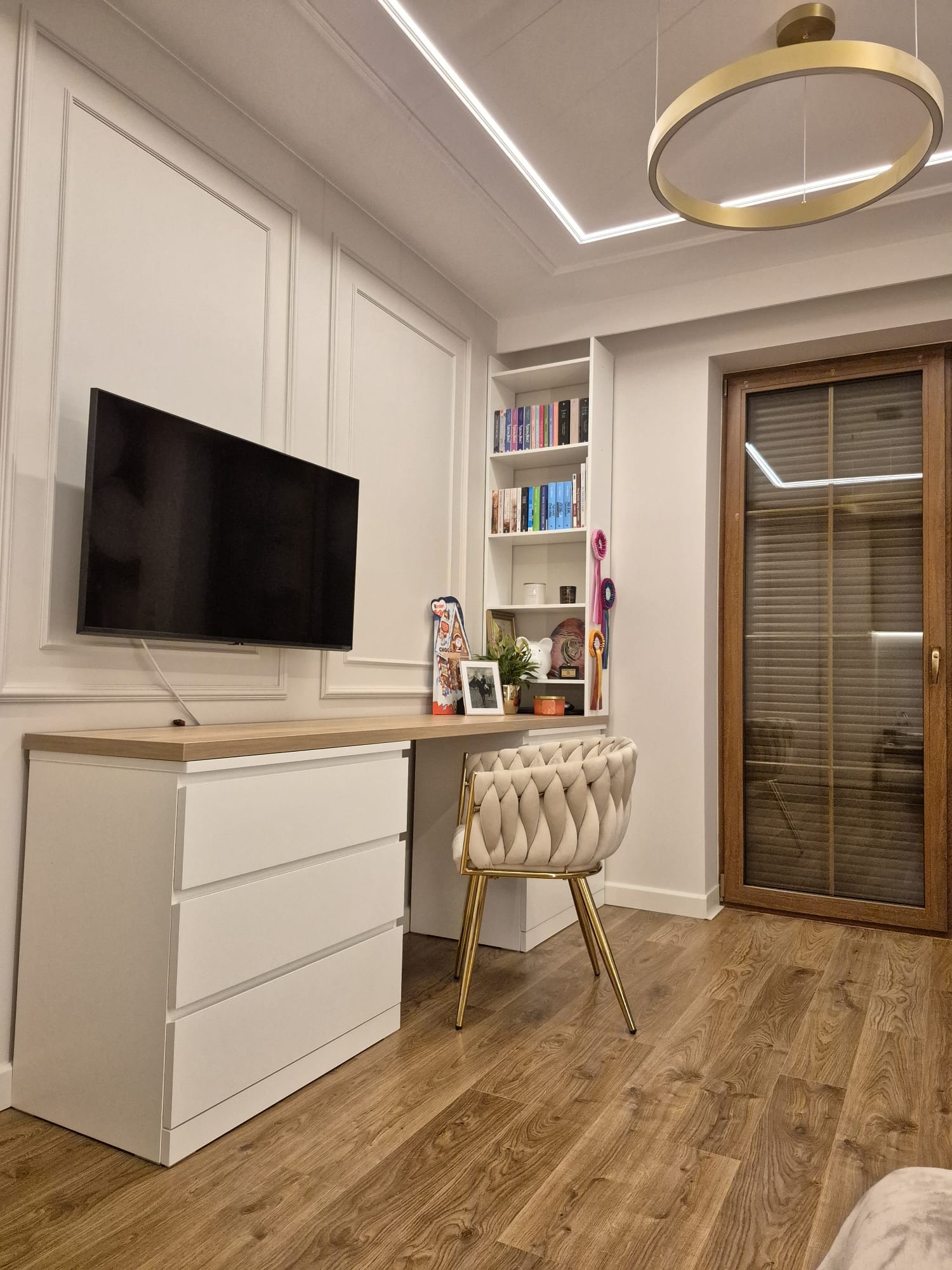 Pokój po wykończeniu: białe ściany z dekoracyjnymi listwami, drewniana podłoga, biurko z komodą, telewizor na ścianie, okno z żaluzjami i designerskie oświetlenie.