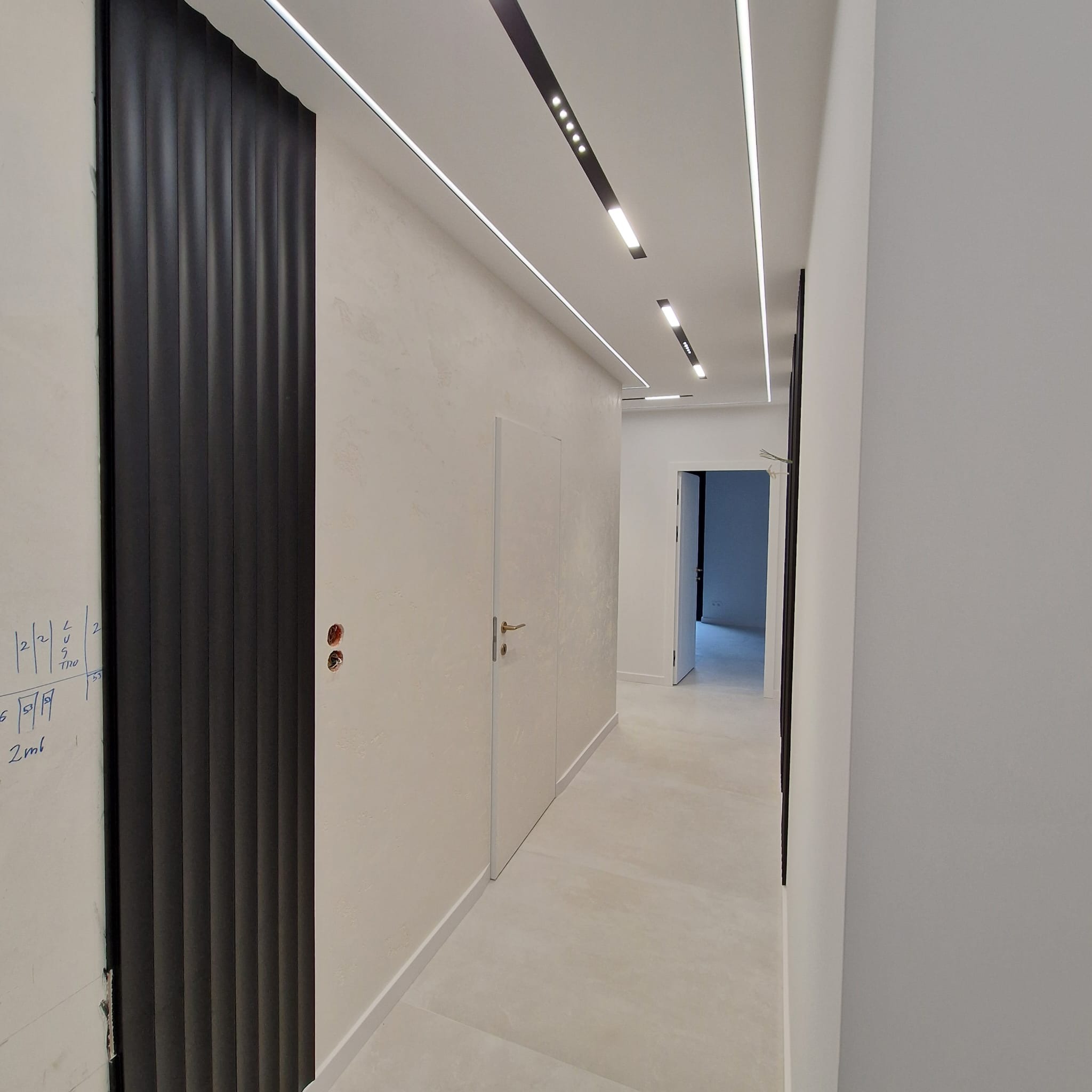 Nowoczesny korytarz po remoncie: białe ściany z fakturą, ukryte drzwi, czarna lamelowa ściana i liniowe oświetlenie sufitowe LED. Minimalistyczny design.