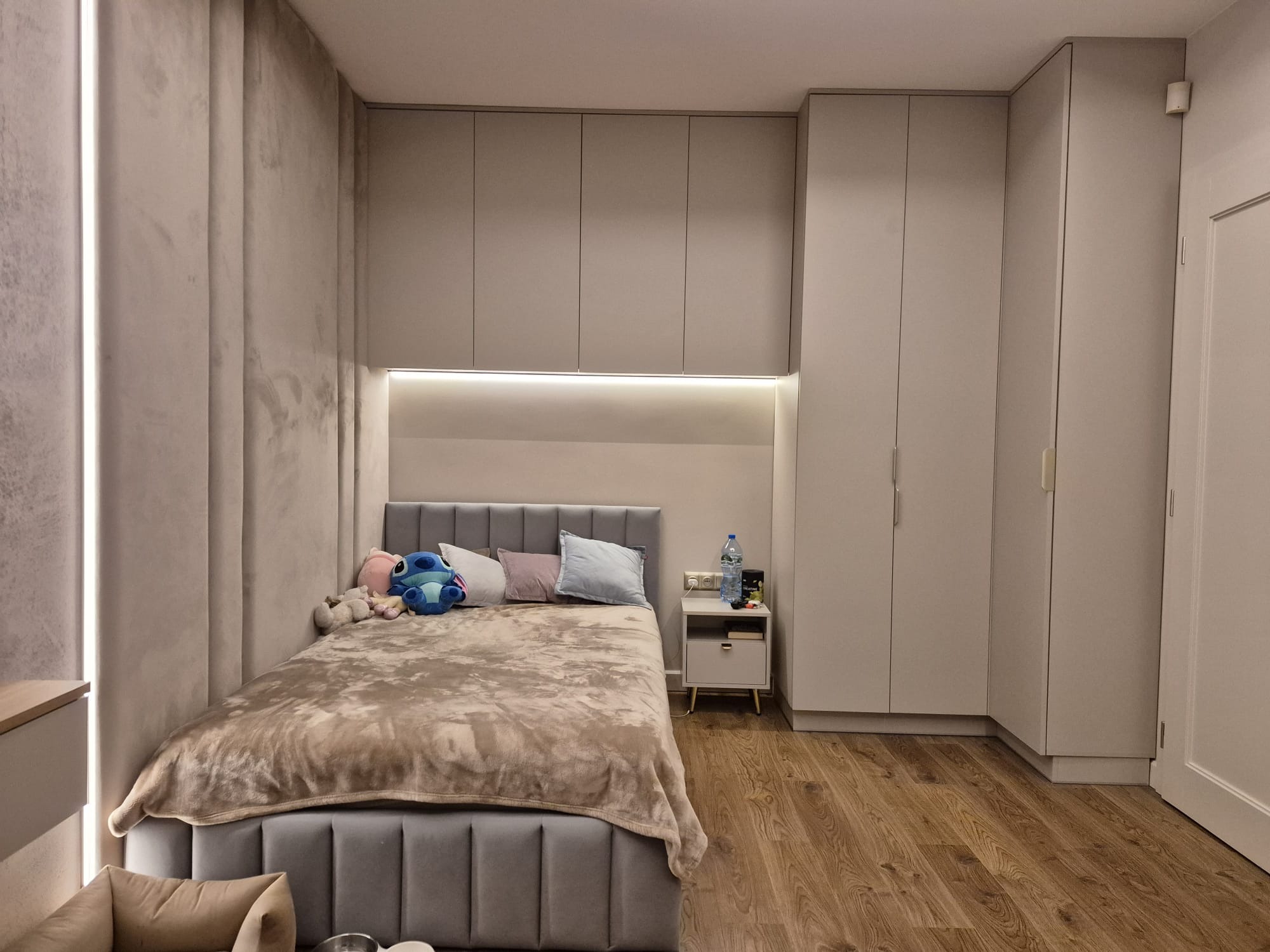 Nowoczesna sypialnia w stonowanych barwach: szara tapeta z efektem betonu, łóżko z tapicerowanym zagłówkiem, drewniana podłoga i zabudowa meblowa pod sufit.