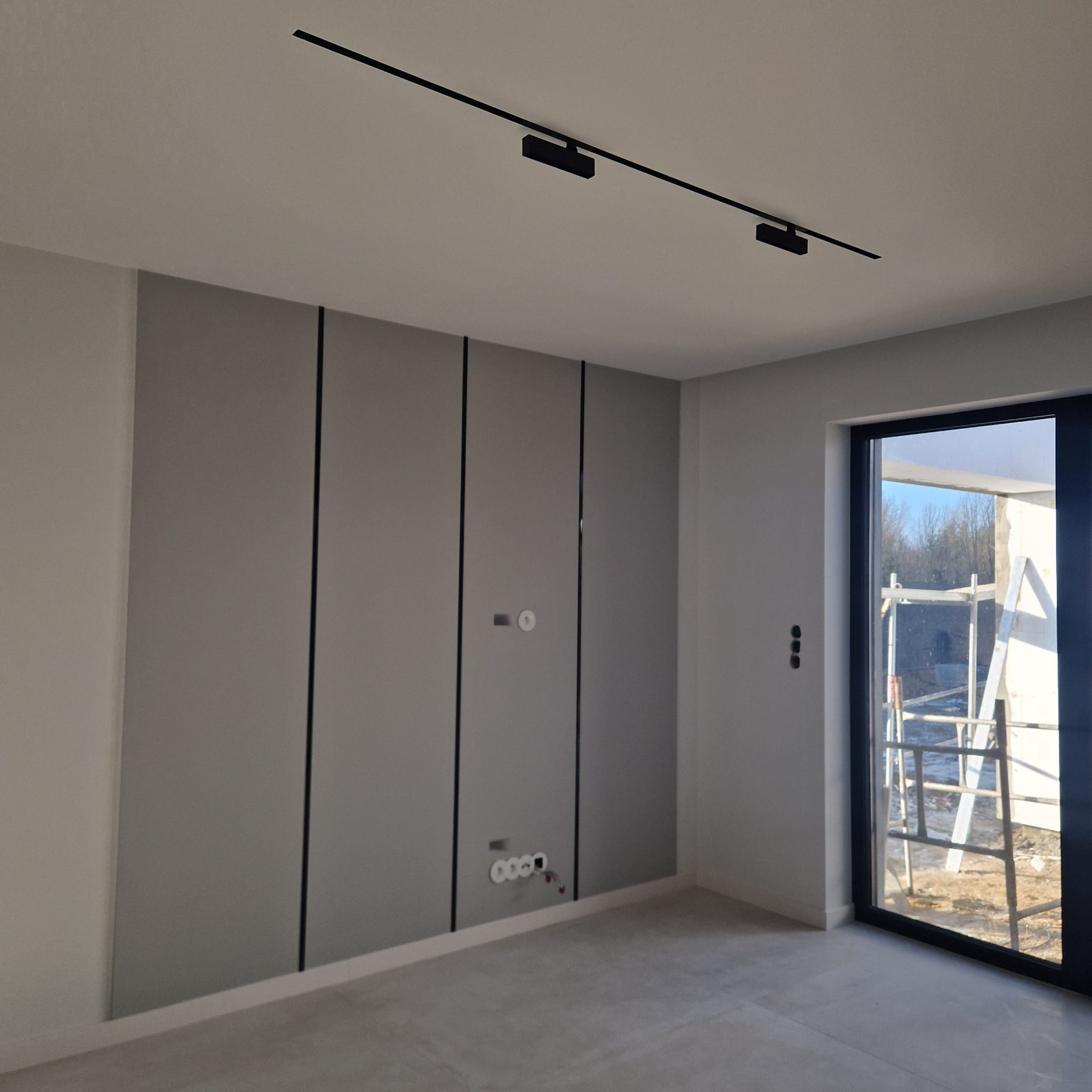 Szare panele ścienne z czarnymi listwami w nowym wnętrzu. Widoczne gniazdka elektryczne i czarna listwa oświetleniowa na suficie. Za oknem rusztowanie.