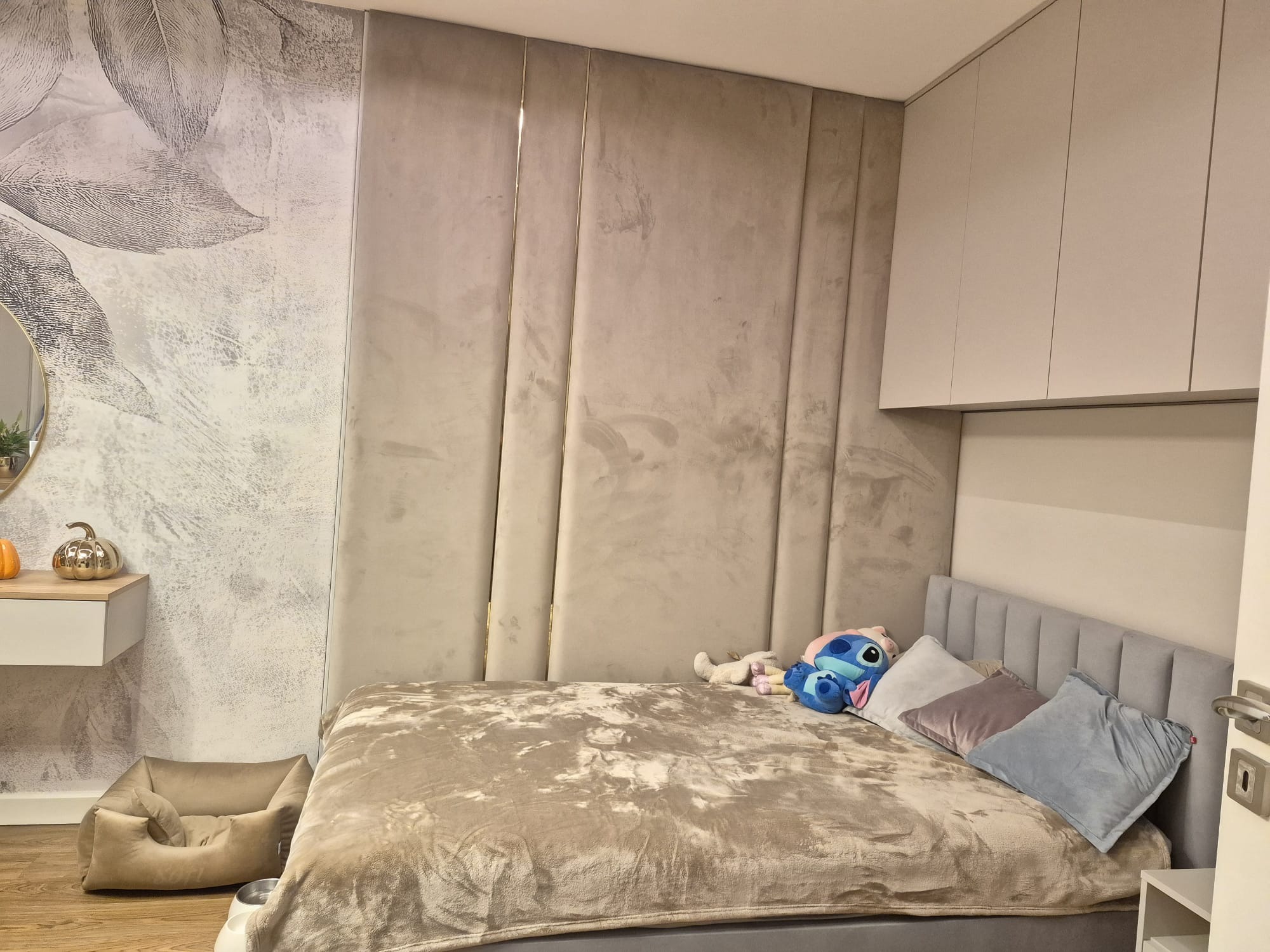 Beżowa sypialnia z tapetą w liście, pluszowym łóżkiem i panelem ściennym. Uwagę zwraca designerska lampka nocna i legowisko dla psa w rogu.