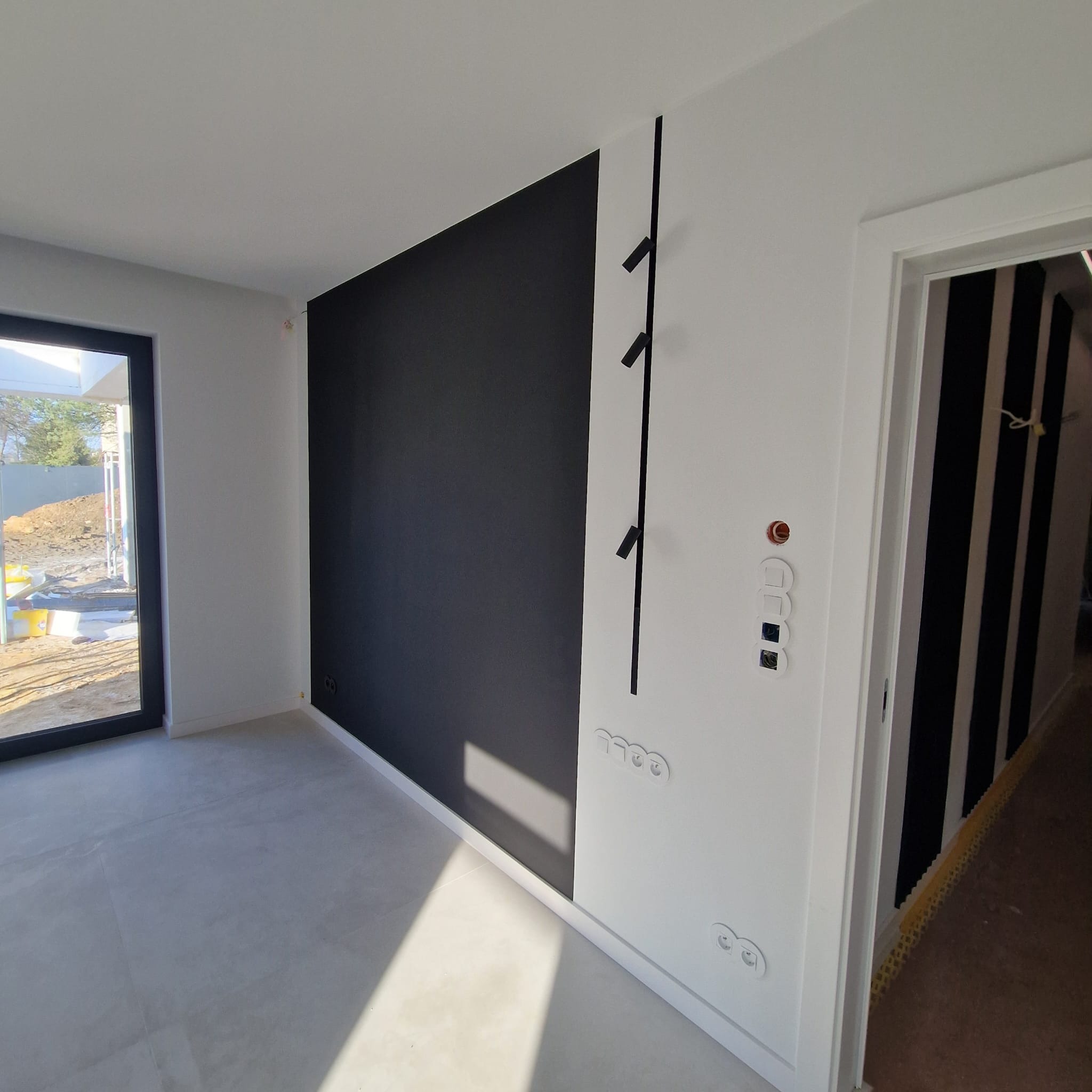 Minimalistyczne wnętrze: czarno-biała ściana z wieszakiem, widoczne gniazdka i włączniki. W tle drzwi z pionowymi pasami i okno z widokiem na plac budowy.