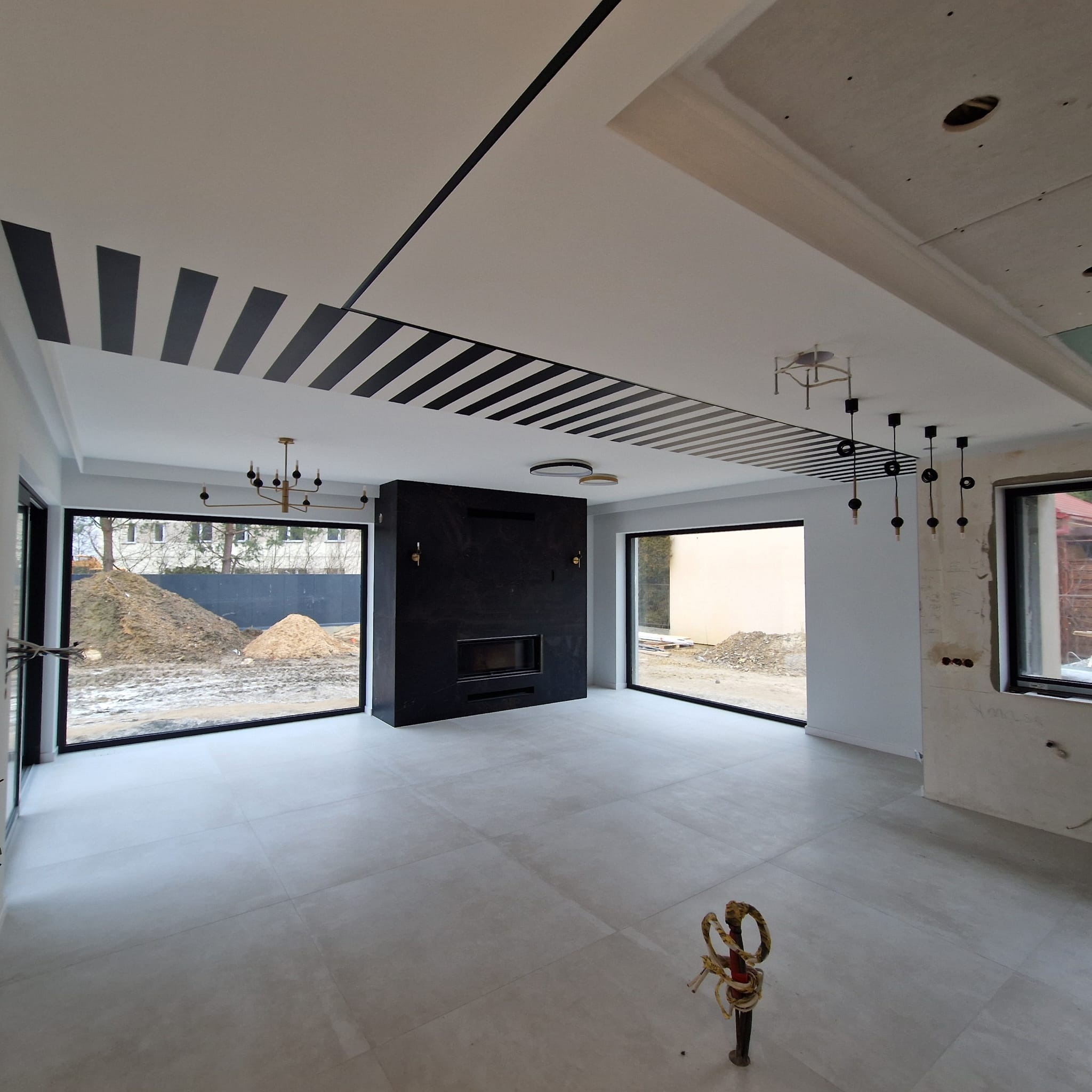 Przestronne wnętrze z nowoczesnym kominkiem i wzorzystym sufitem w czarno-białe pasy. Duże okna z widokiem na plac budowy. Minimalistyczne lampy wiszące.