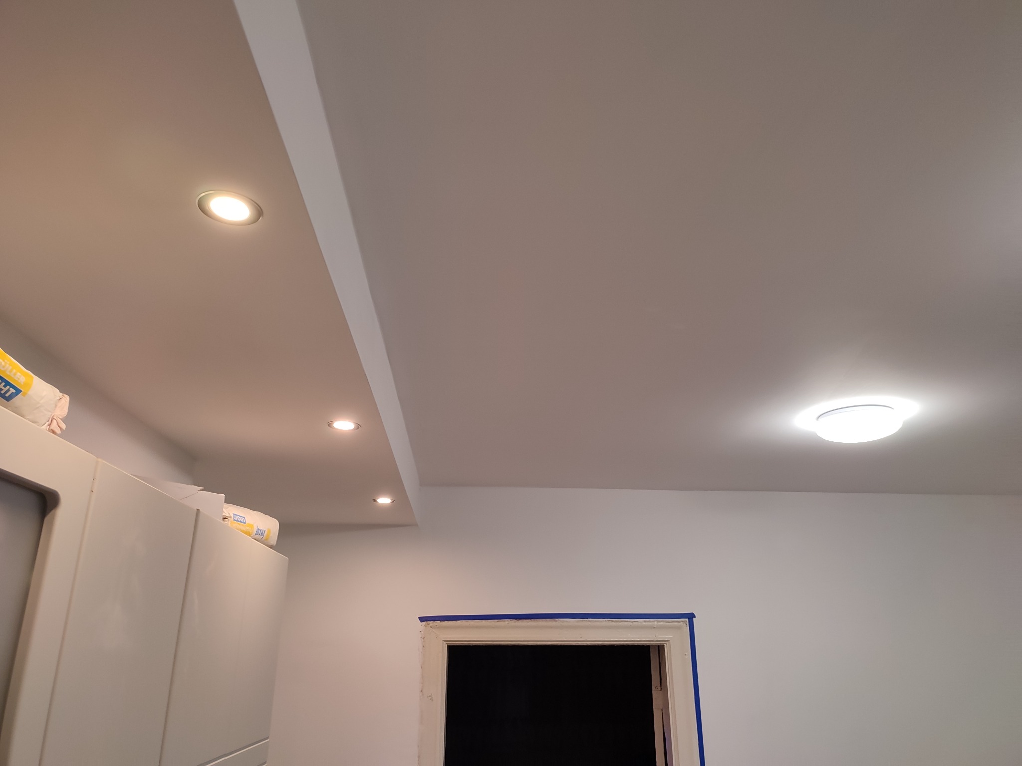 Wykończone wnętrze z gładkimi, pomalowanymi ścianami i sufitem, z zamontowanymi punktowymi lampami LED oraz przygotowanym do dalszych prac otworem drzwiowym zabezpieczonym niebieską taśmą malarską.