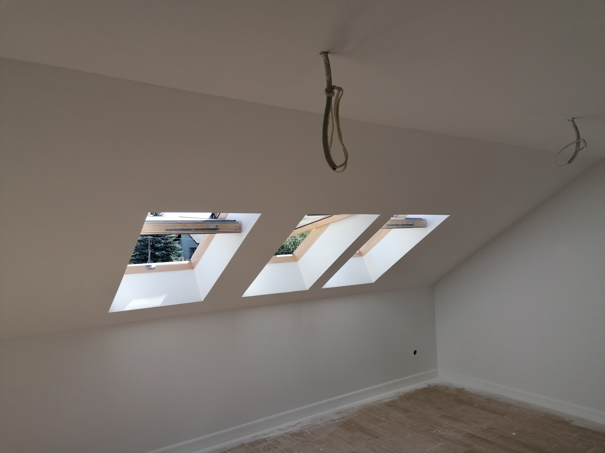 Poddasze po remoncie z trzema oknami dachowymi, świeżo pomalowane ściany na biało, widoczne przewody elektryczne zwisające z sufitu.