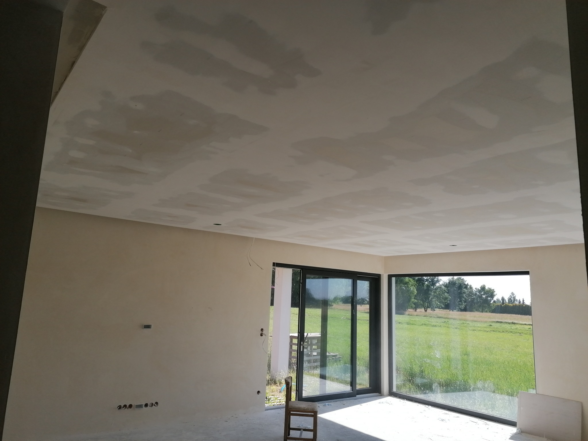 Wnętrze w trakcie prac wykończeniowych: sufit z widocznymi łączeniami płyt, ściany przygotowane pod malowanie, duże okna z widokiem na zieleń, krzesło w rogu pomieszczenia.