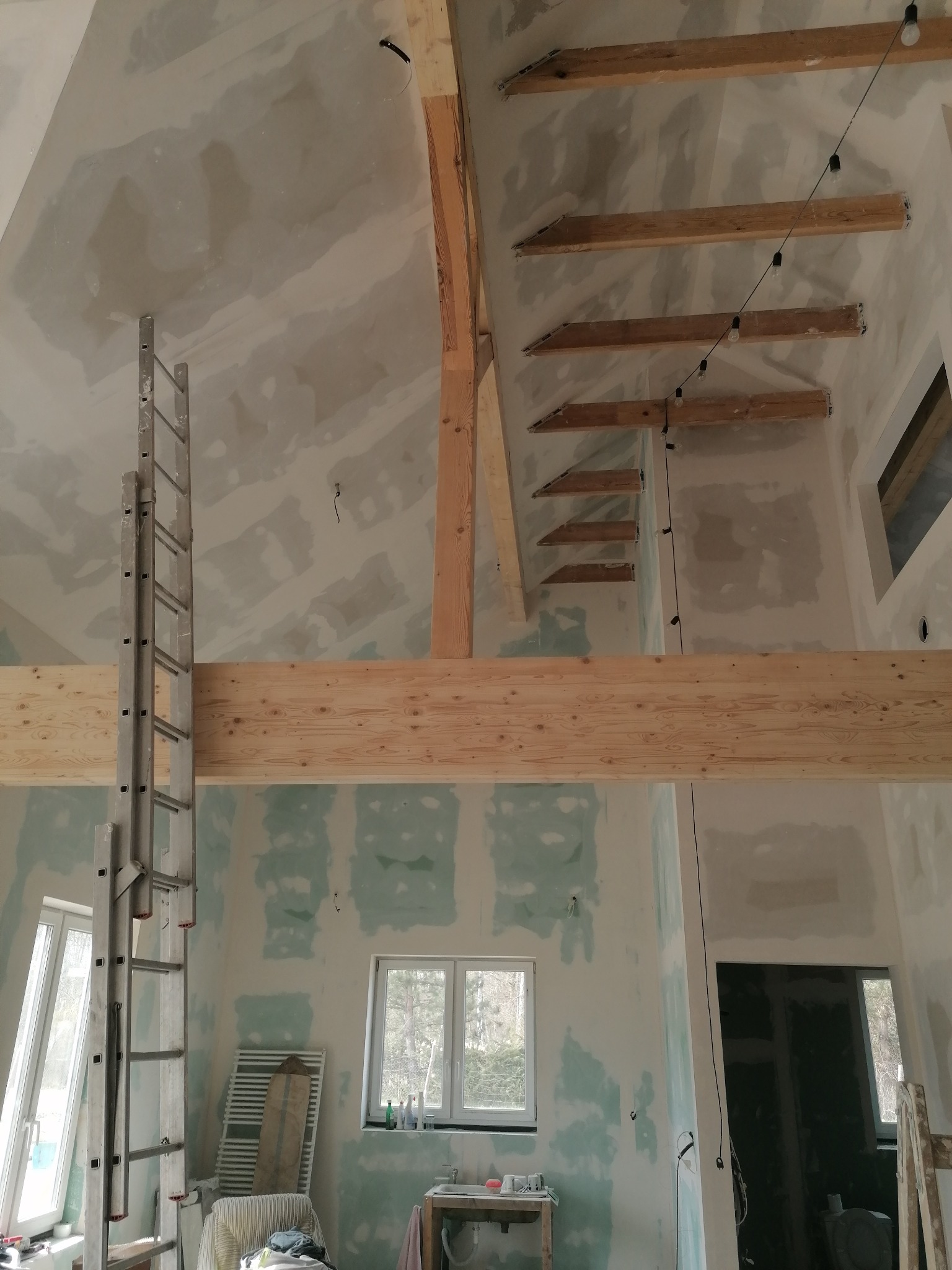 Poddasze w trakcie remontu: ściany i sufit pokryte płytami karton-gips z widocznymi łączeniami, drewniane belki konstrukcyjne, aluminiowa drabina oparta o ścianę, okna z widokiem na zewnątrz.