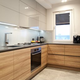 STUDIO PROJEKTOWE Kampra Meble - Nowoczesna kuchnia w kształcie litery L z drewnianymi frontami szafek dolnych i białymi, lakierowanymi szafkami górnymi, szary blat i płytki podłogowe, oświetlenie LED pod szafkami.