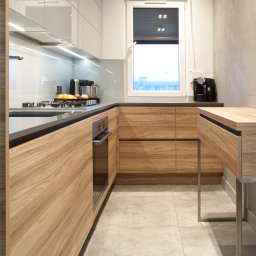 STUDIO PROJEKTOWE Kampra Meble - Wąska kuchnia z drewnianymi frontami szafek, czarnym blatem i szarą glazurą, oświetlona światłem dziennym wpadającym przez okno.