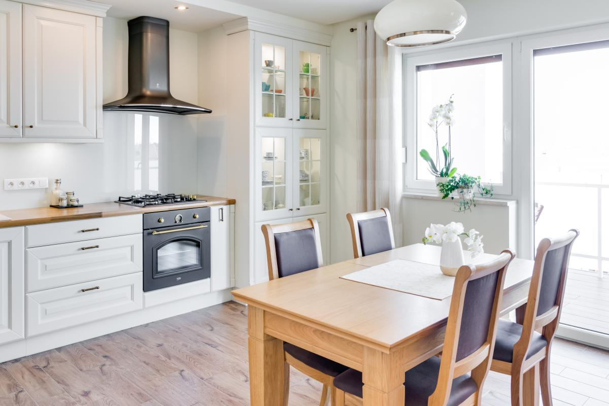 Jasna kuchnia z drewnianym blatem, czarnym okapem i piekarnikiem w stylu retro. Stół jadalniany z krzesłami w pobliżu okna z kwiatami.