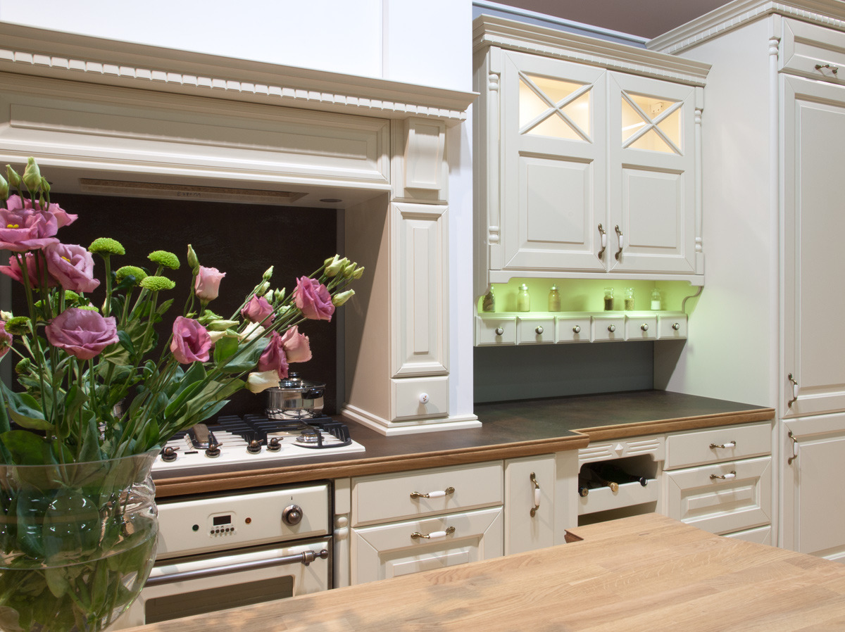 Elegancka kuchnia w stylu retro z kremowymi szafkami, drewnianym blatem i bukietem różowych kwiatów w wazonie na pierwszym planie, z widoczną płytą grzewczą i piekarnikiem z detalami w stylu vintage.
