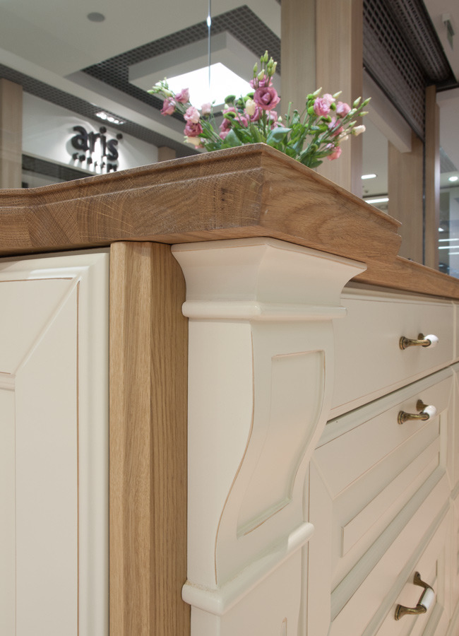 Fragment klasycznej kuchni: drewniany blat, kremowe fronty szafek z dekoracyjnymi uchwytami i kwiatami w tle, widok na wnętrze salonu meblowego.